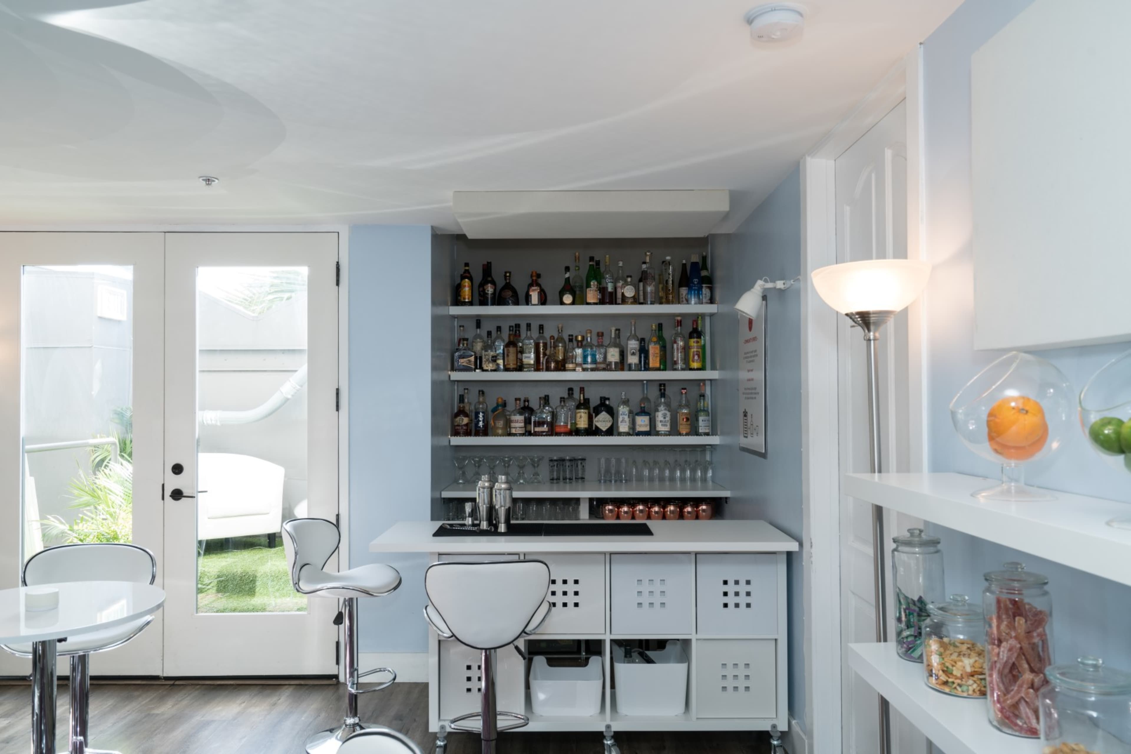 A modern bar area with a white counter, high stools, and shelves lined with various bottles, alongside a glass door leading to an outdoor space.