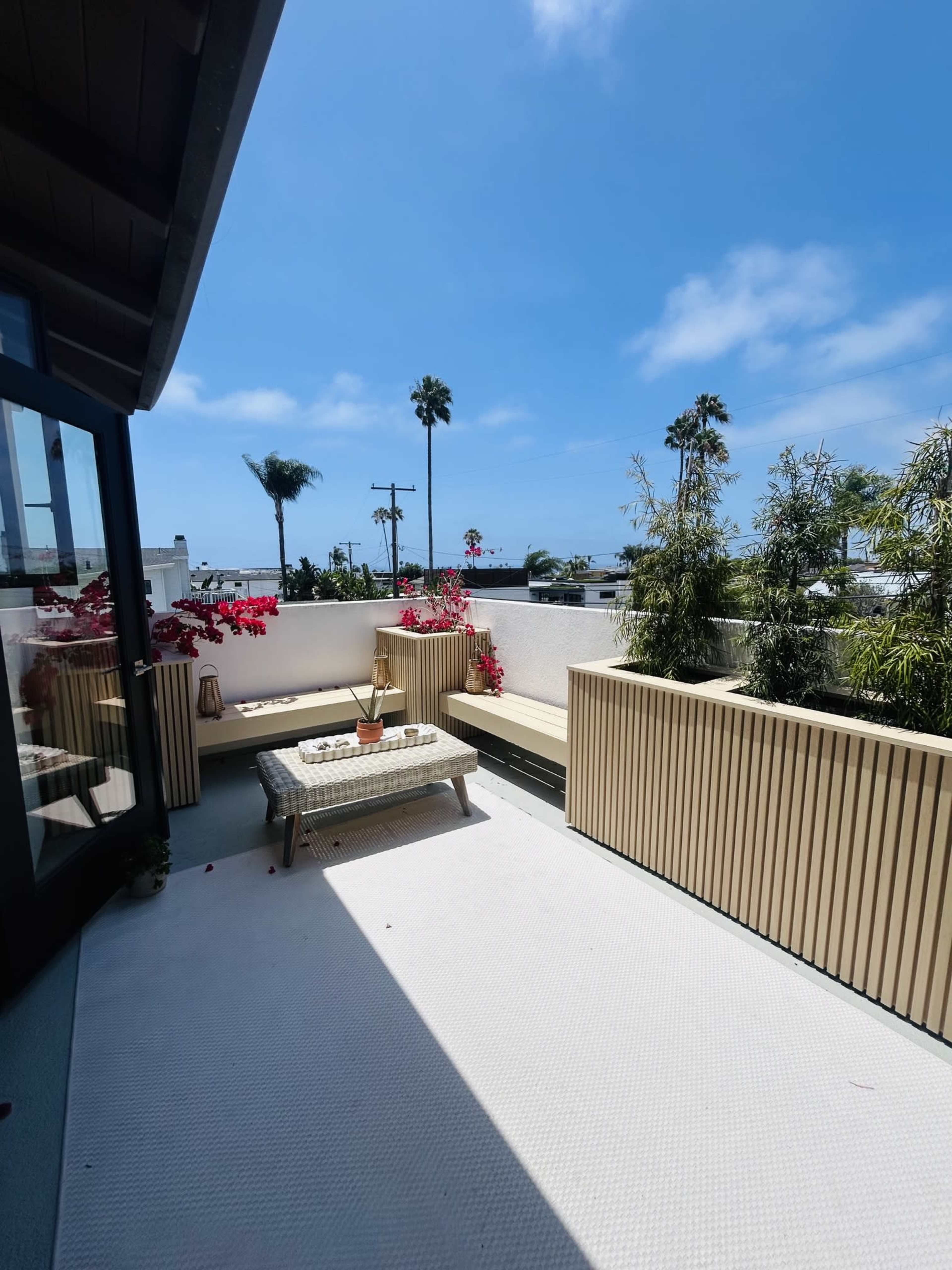 A rooftop terrace features a wicker seating area surrounded by flowering plants and palm trees, with a clear blue sky above.