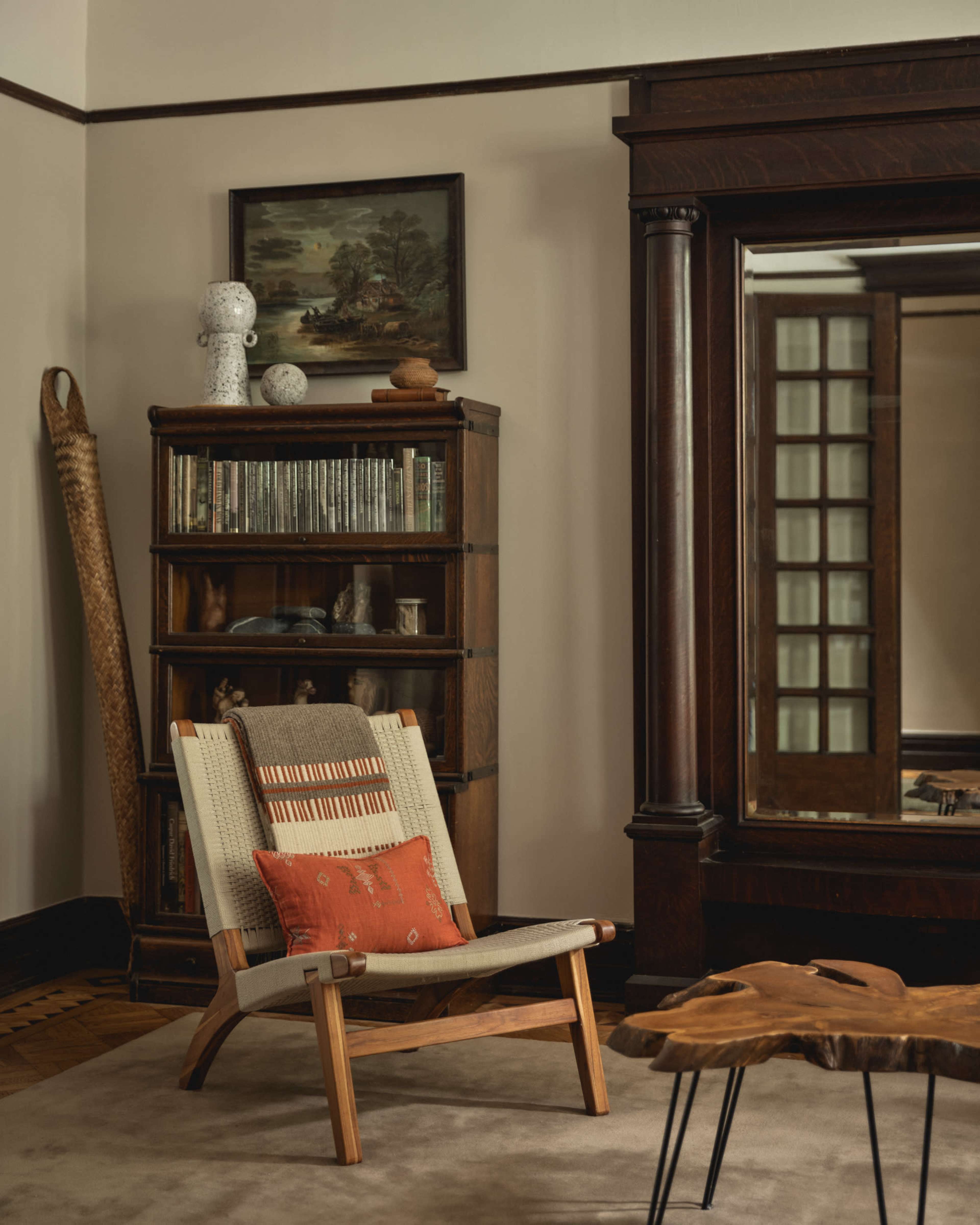 The scene features a wooden chair with a patterned cushion positioned beside a bookshelf filled with books and decorative items, against a backdrop of an ornate mirror and a neutral carpet.