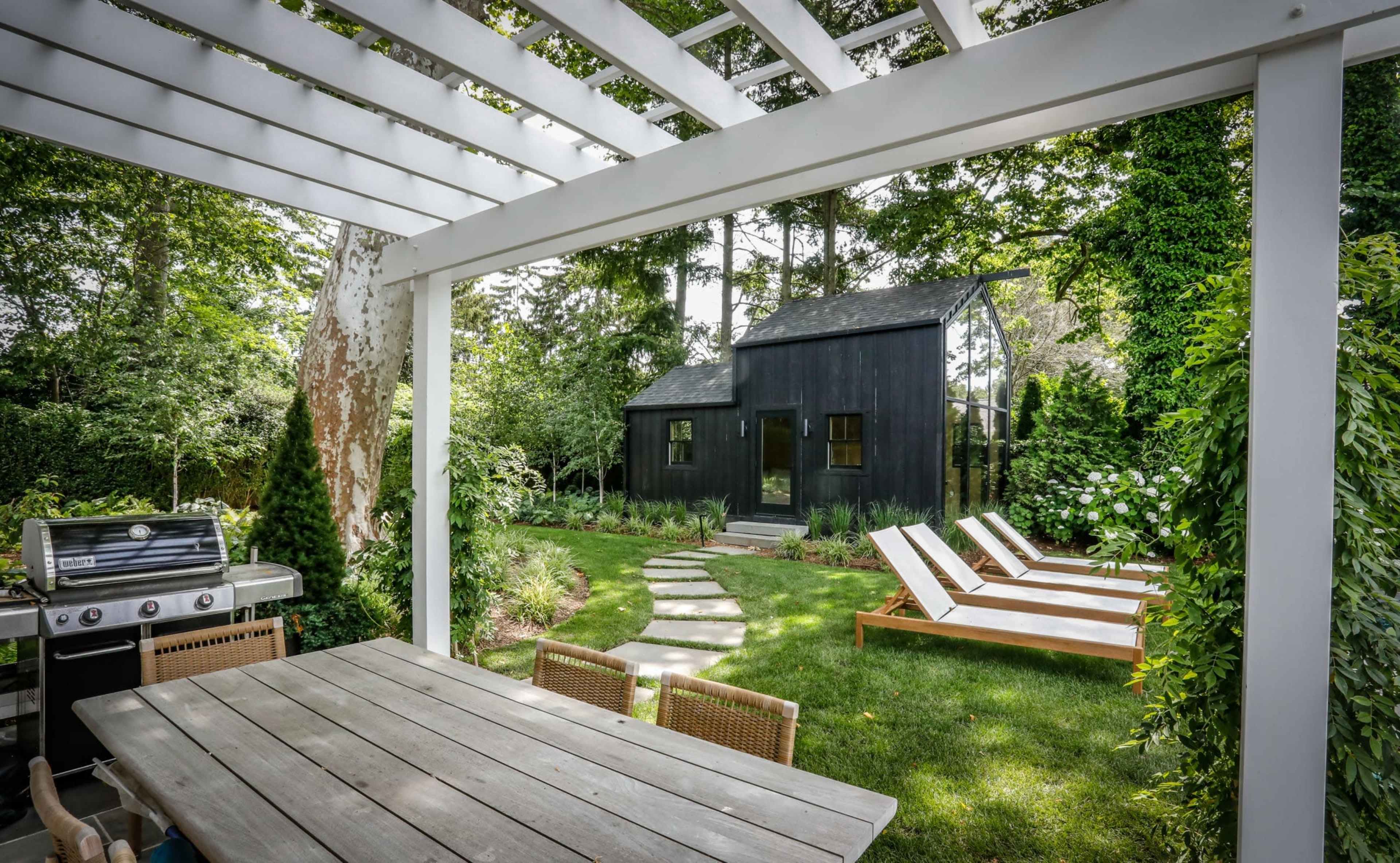 A backyard features a wooden dining table, a barbecue grill, and white lounge chairs set along a stone path leading to a small black cabin.