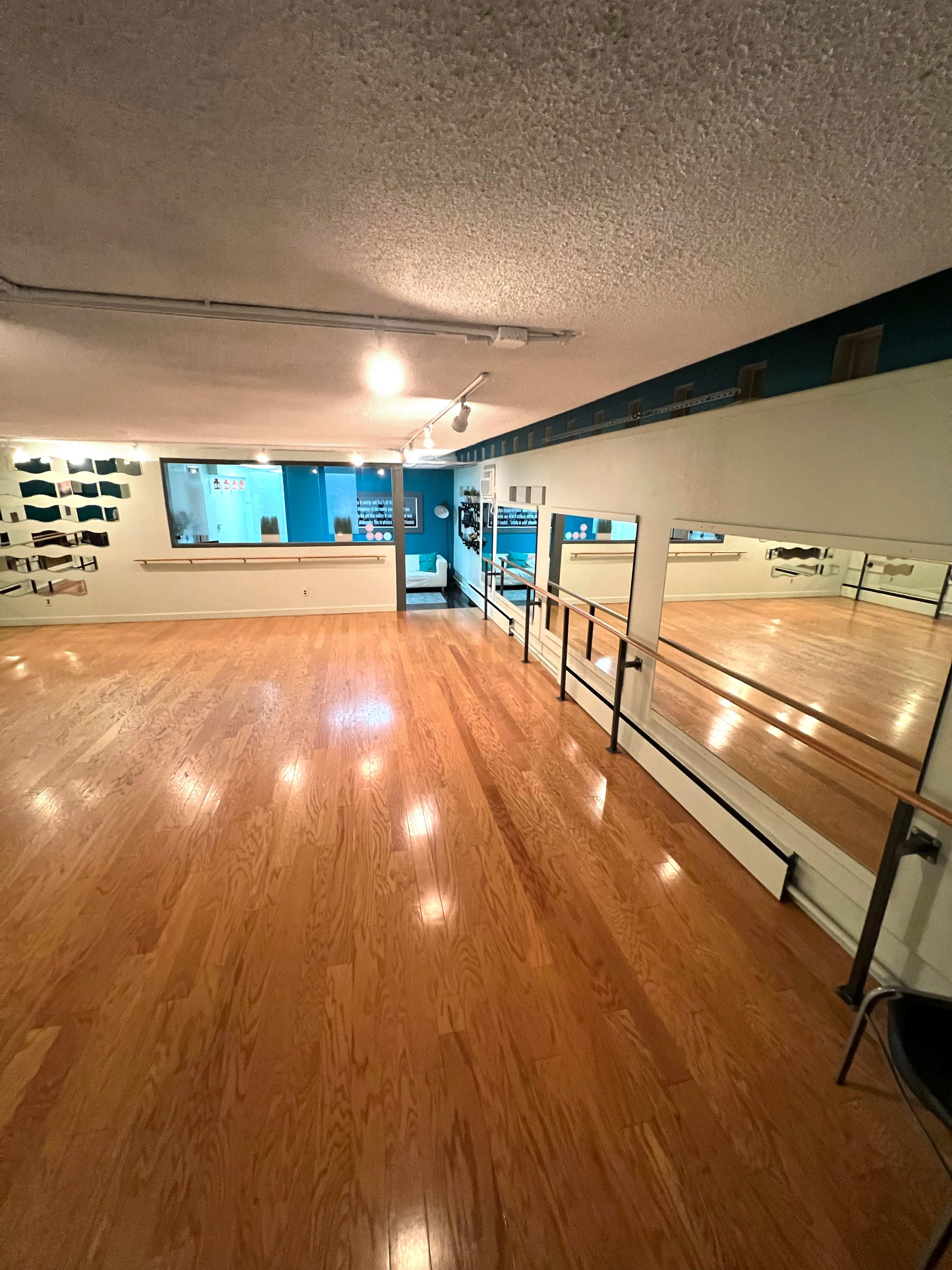 A spacious dance studio with wooden floors and mirrors along one wall, illuminated by overhead lighting.