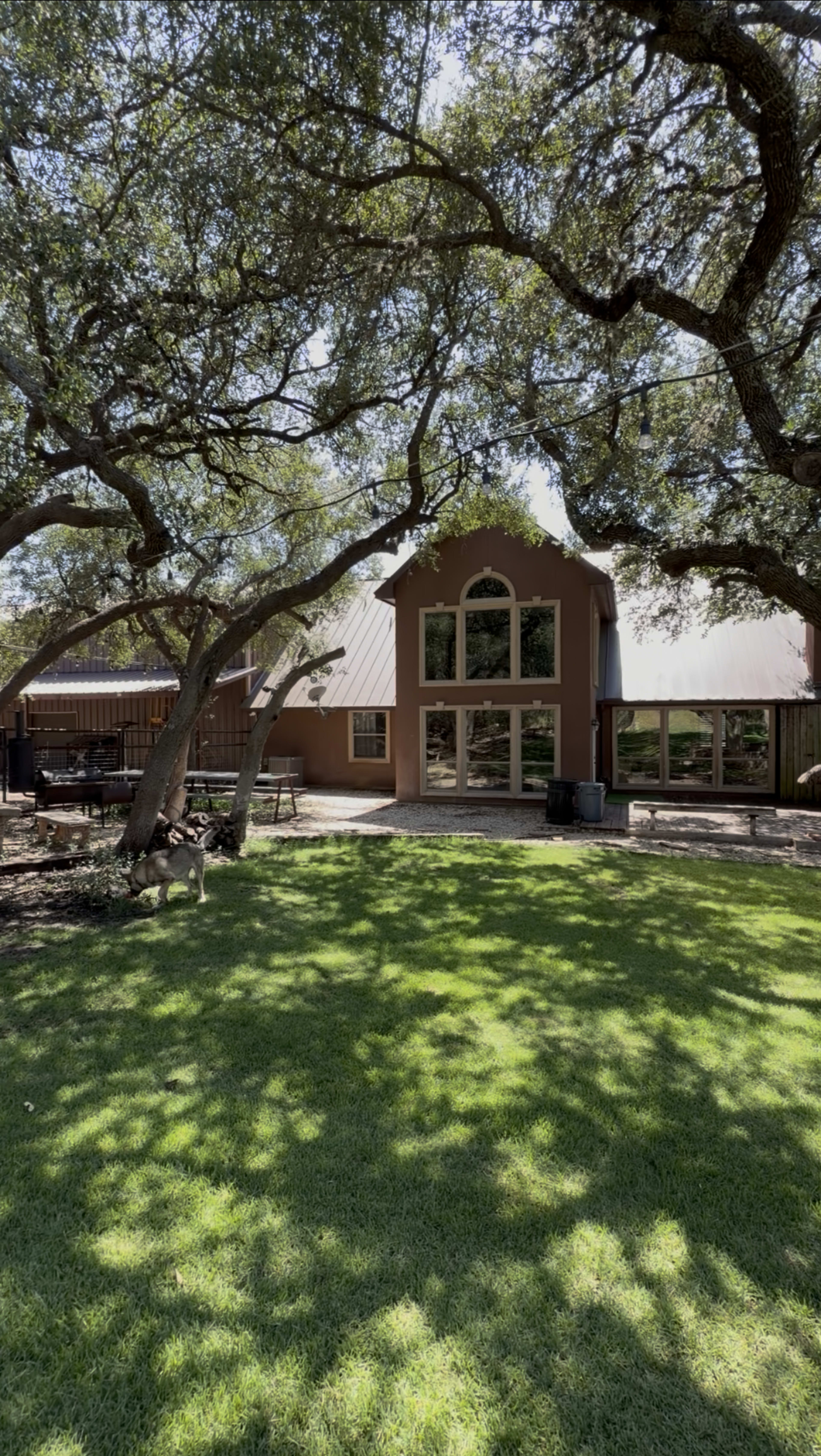 A house with large windows and a gabled roof is surrounded by shaded trees and a neatly maintained lawn.