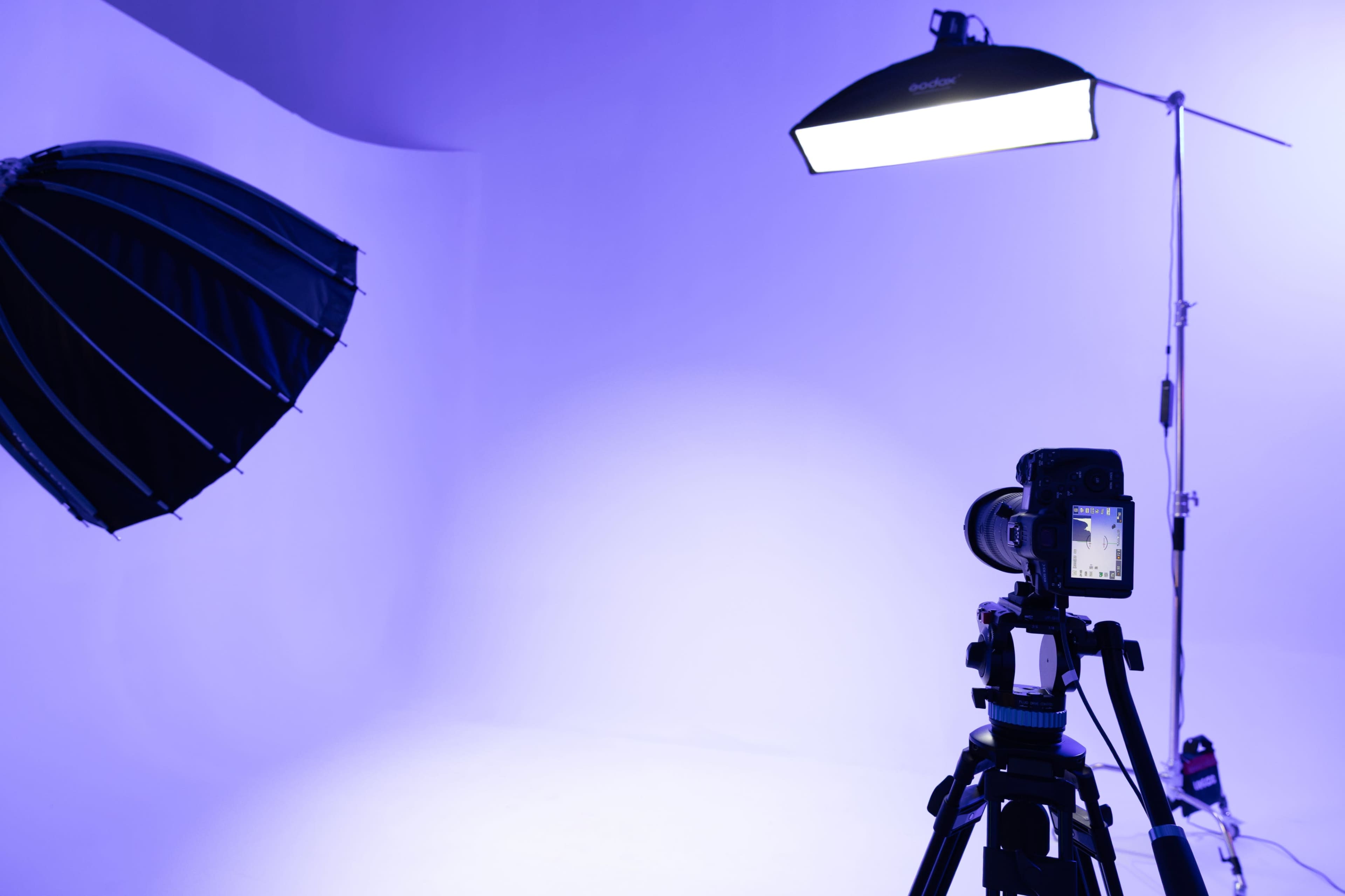 A camera on a tripod is positioned in a photo studio illuminated by an overhead light and a soft umbrella reflector.