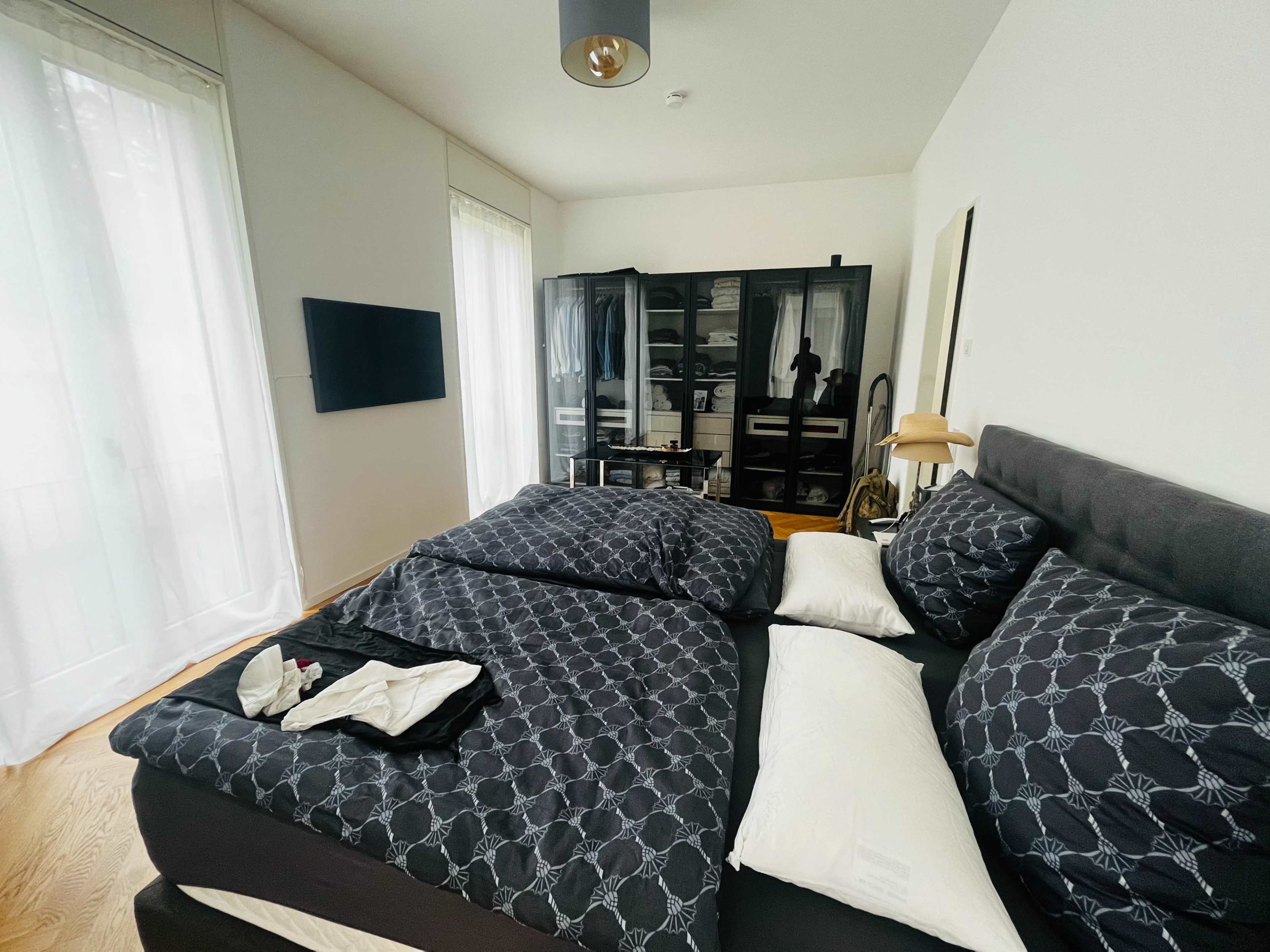 A modern bedroom with a large bed, dark bedding, a wall-mounted television, and a black closet against a light-colored wall.