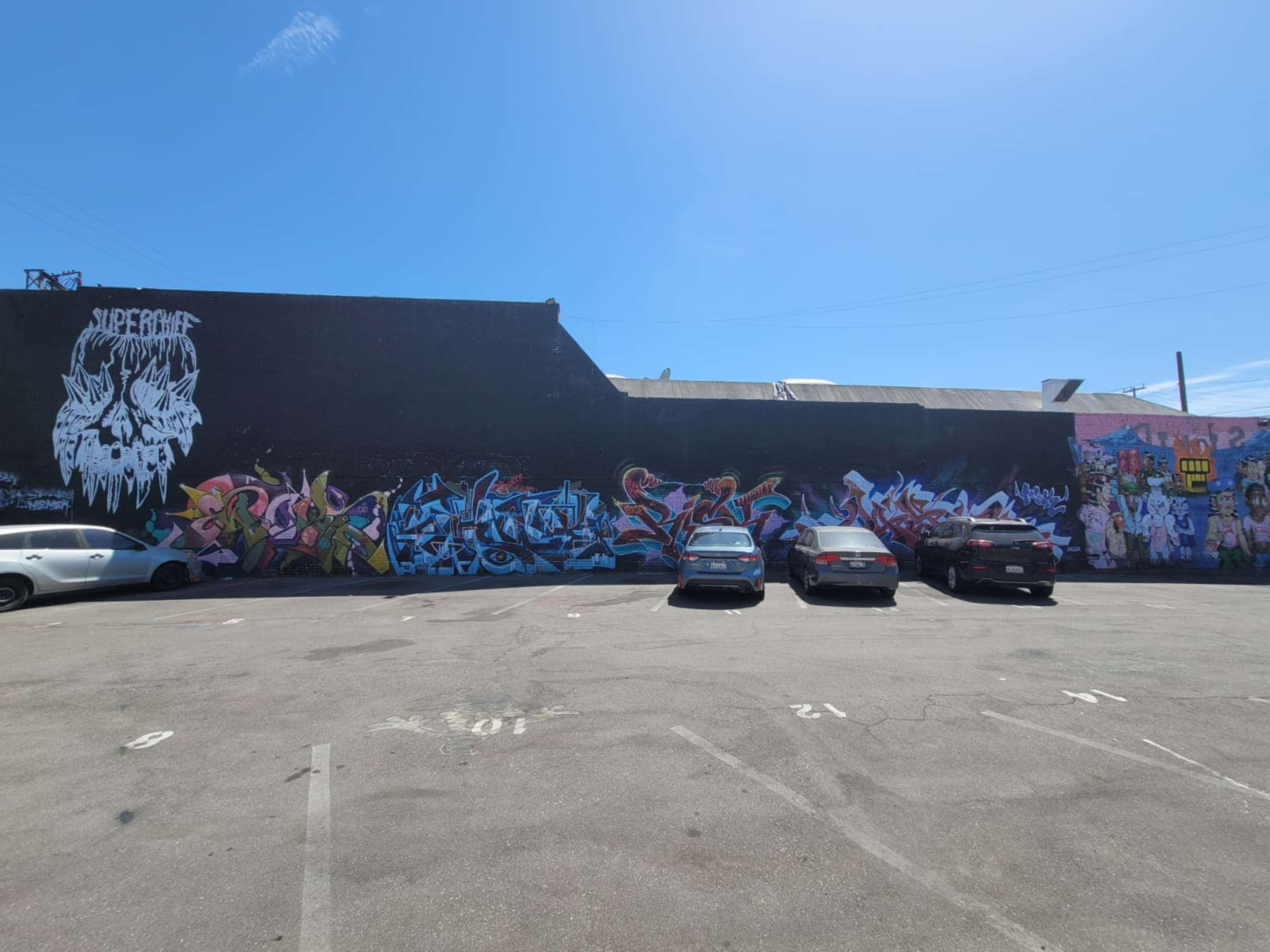 The image shows a long mural covered with colorful graffiti on a black wall, with several parked cars in the foreground.