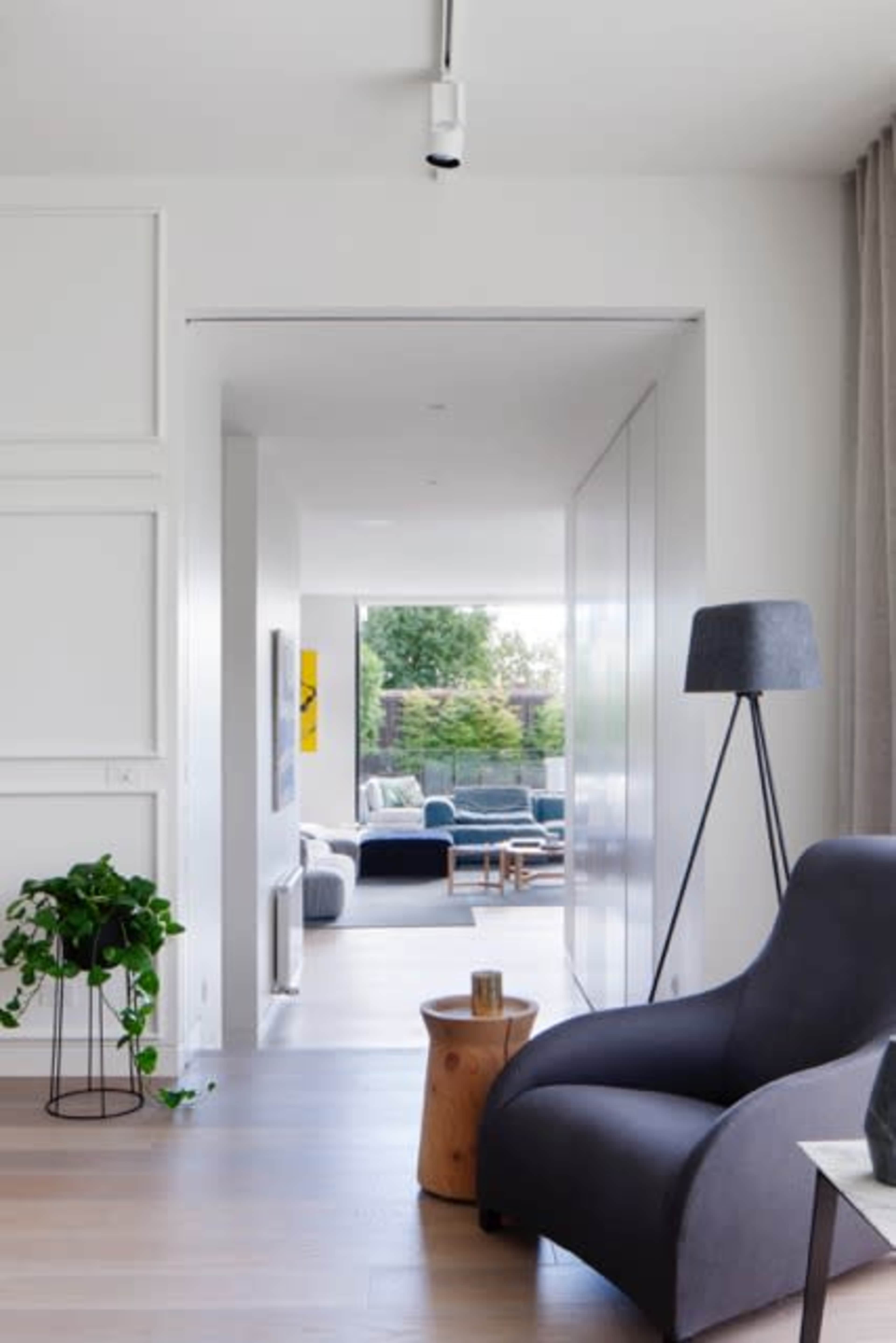 A modern living space features an open layout with a dark armchair, a small wooden side table, and greenery, leading to a sunlit area with couches visible in the background.