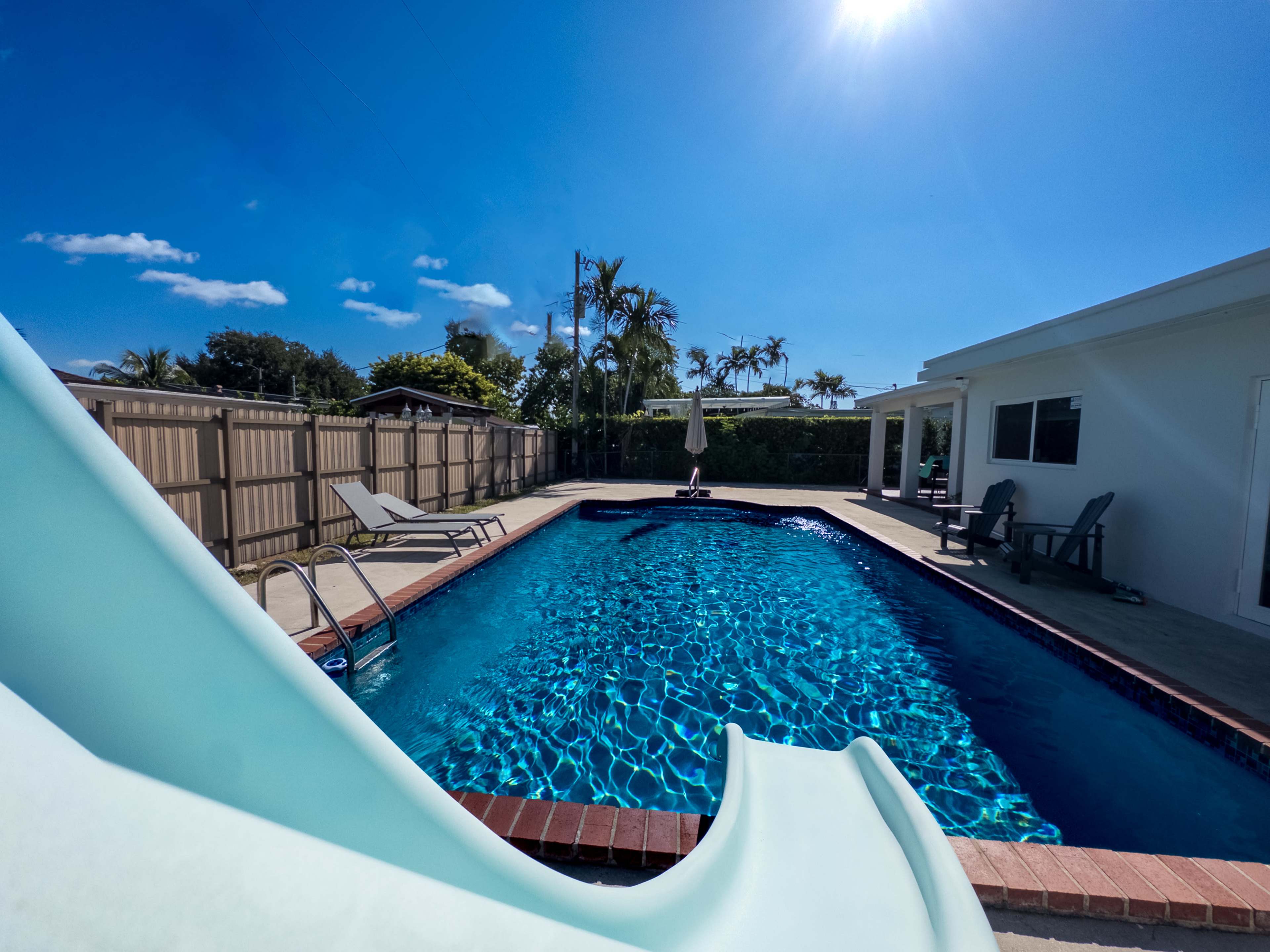 The image shows a clear swimming pool with a slide, surrounded by lounge chairs and a house under a bright blue sky.
