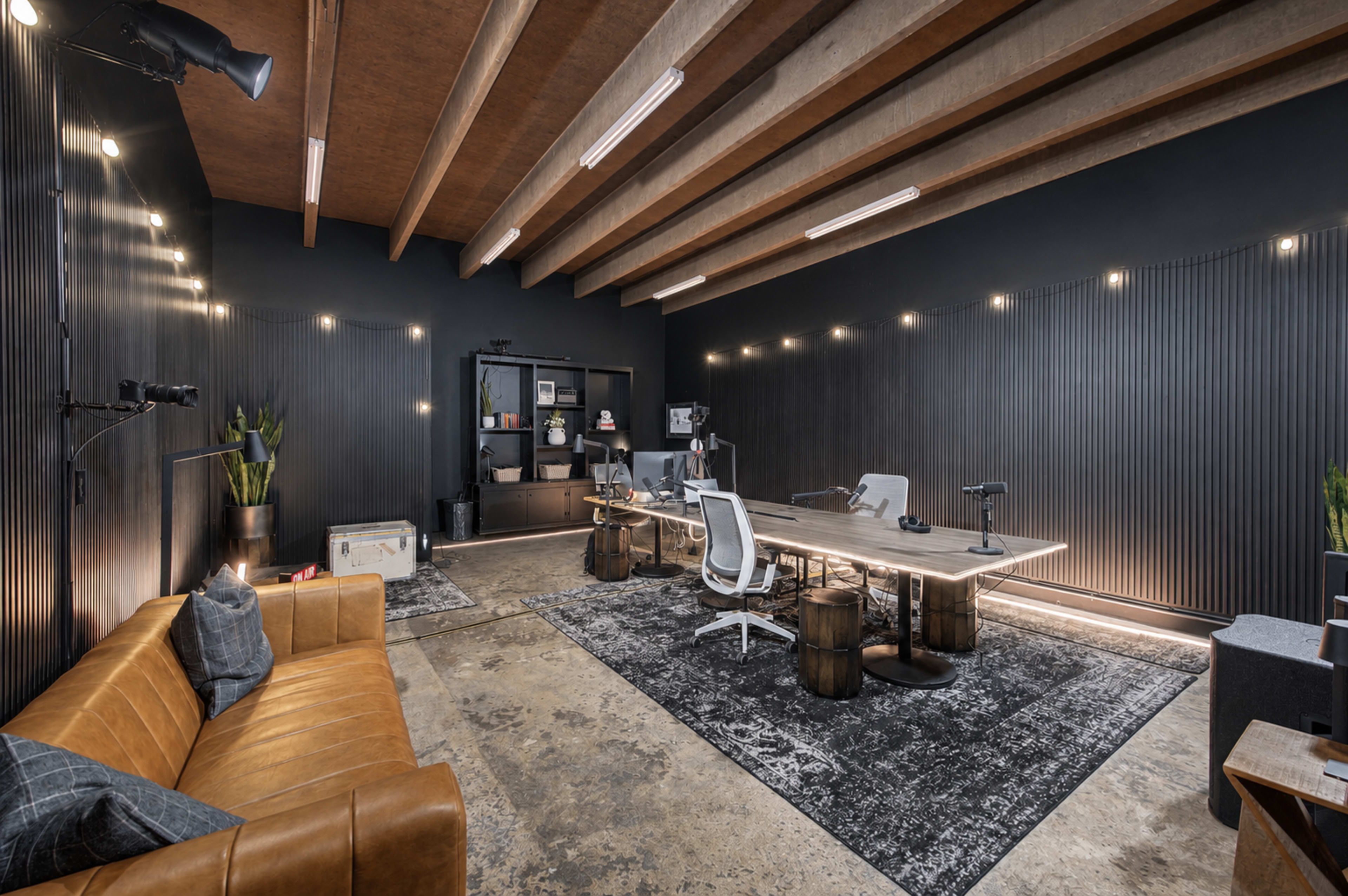The image shows a modern workspace with a large table, several office chairs, and a brown leather sofa, all set against a dark wall with linear wood paneling and ambient lighting.