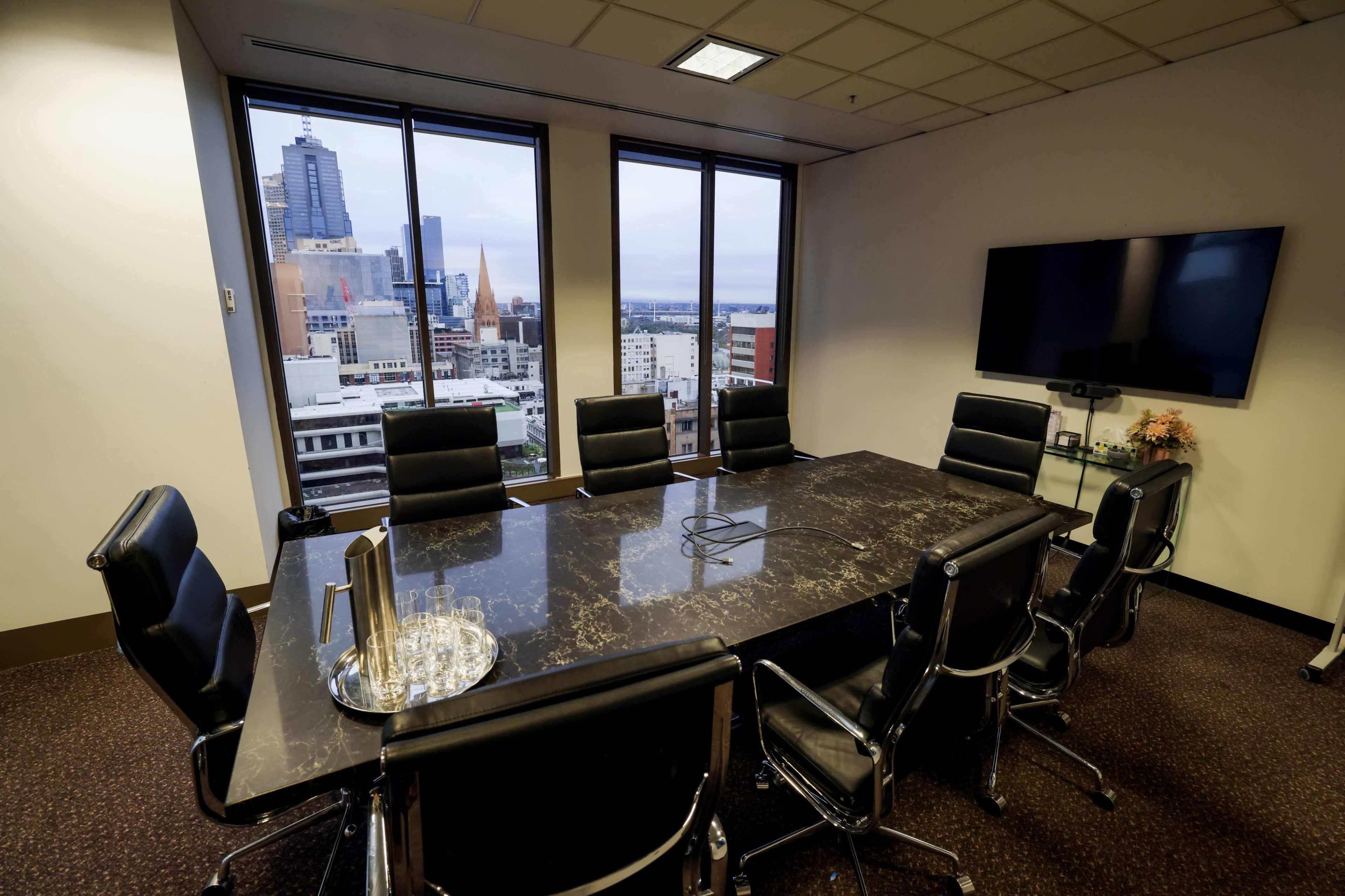 The image shows a modern conference room with a large dark table and black chairs, featuring a city skyline visible through large windows.