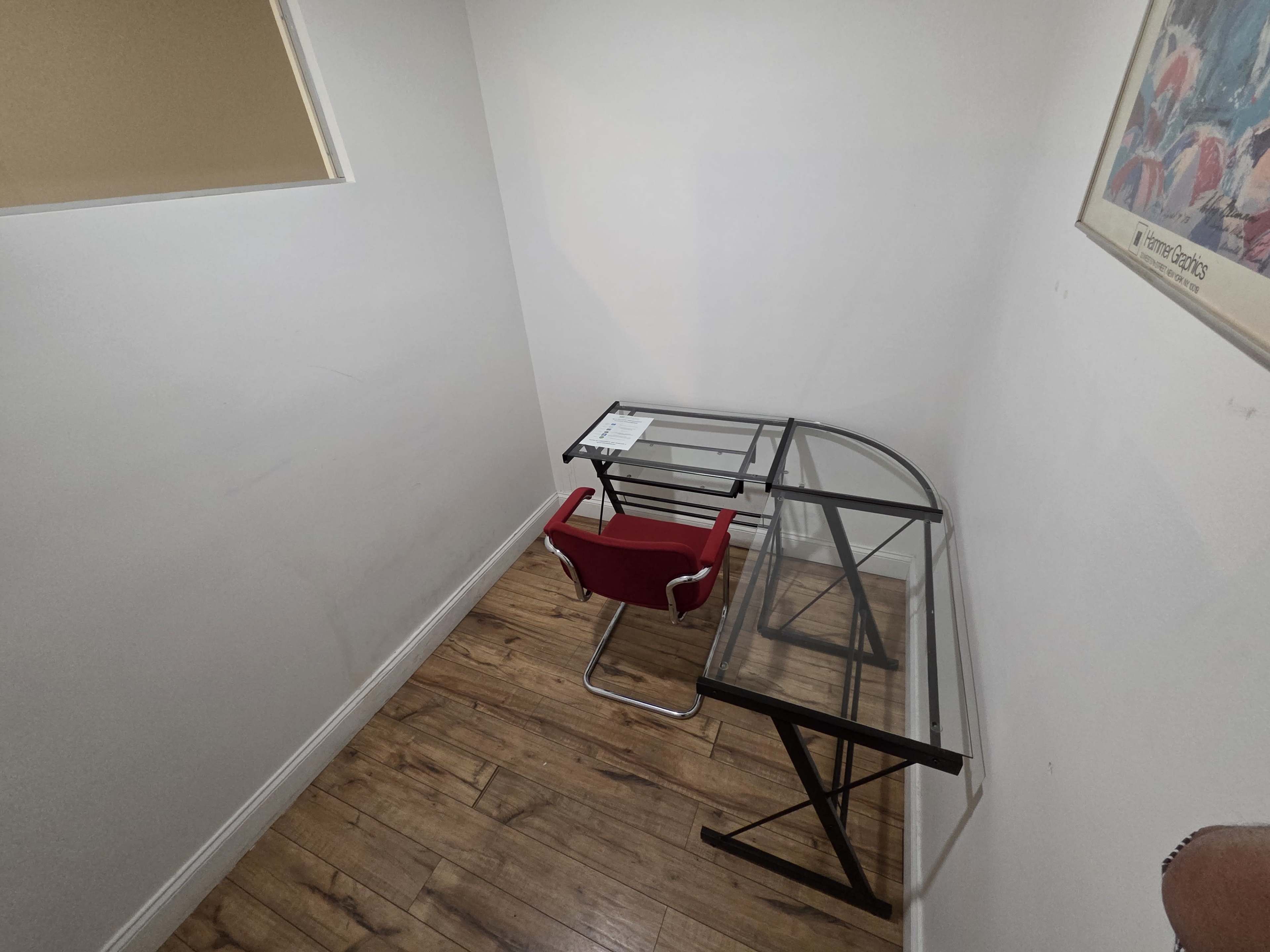 The image shows a small, minimalist office space featuring a glass desk and a red chair against a white wall.