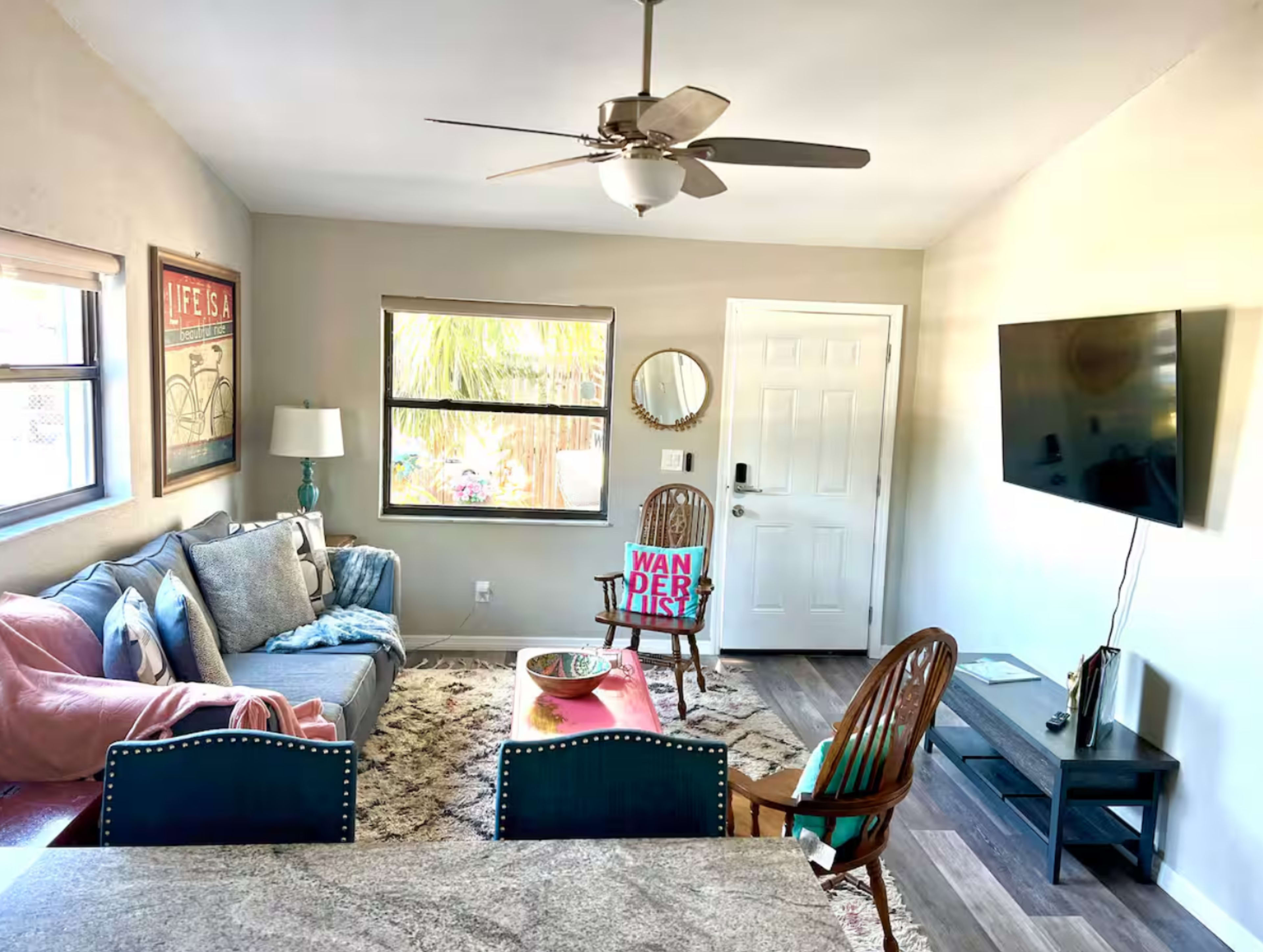 The image shows a cozy living room with a sofa, chair, coffee table, and a mounted television, illuminated by natural light from windows.