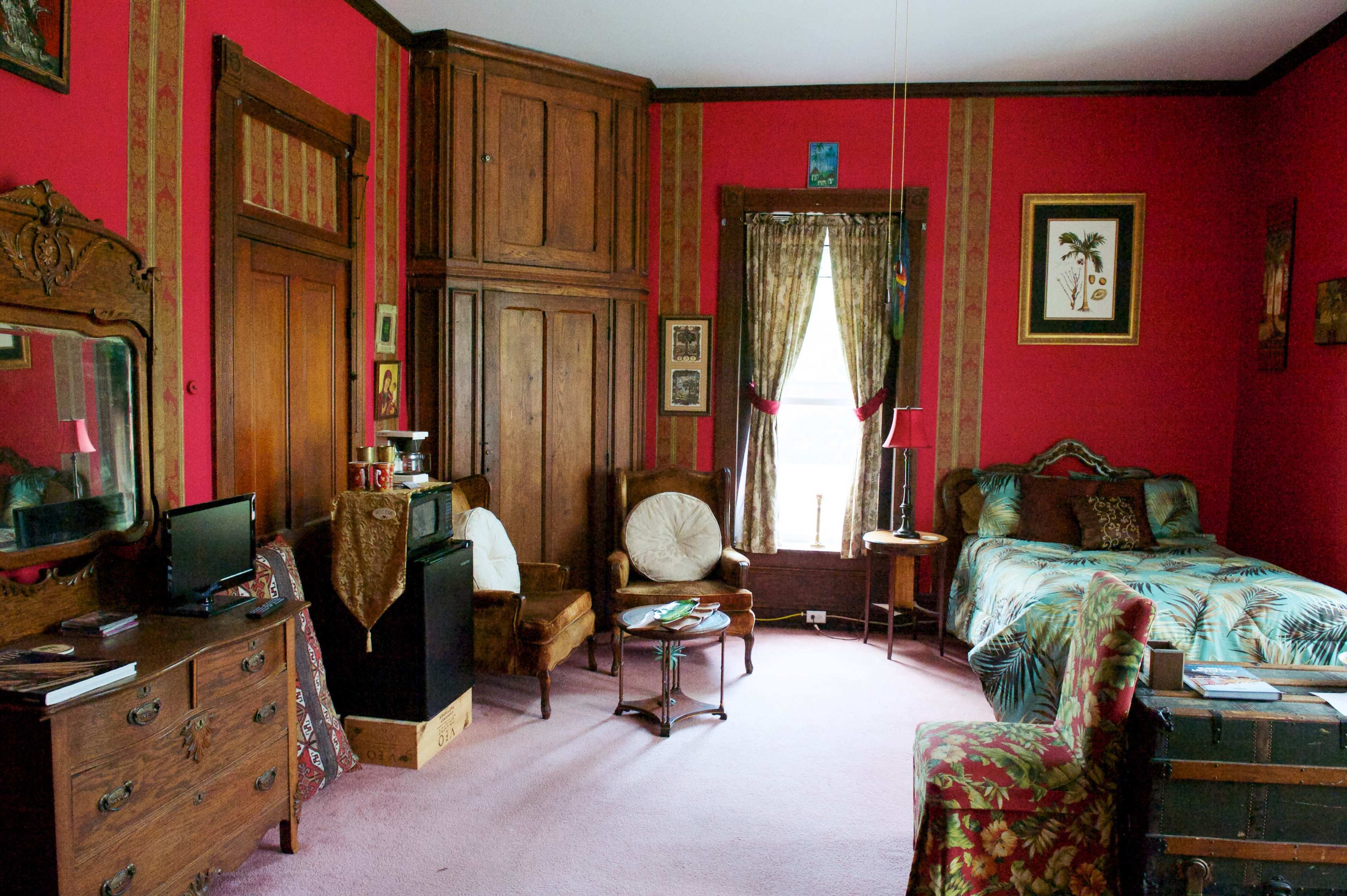 The image shows a cozy bedroom with red walls, a bed with patterned bedding, an antique dresser, and various pieces of wall art.