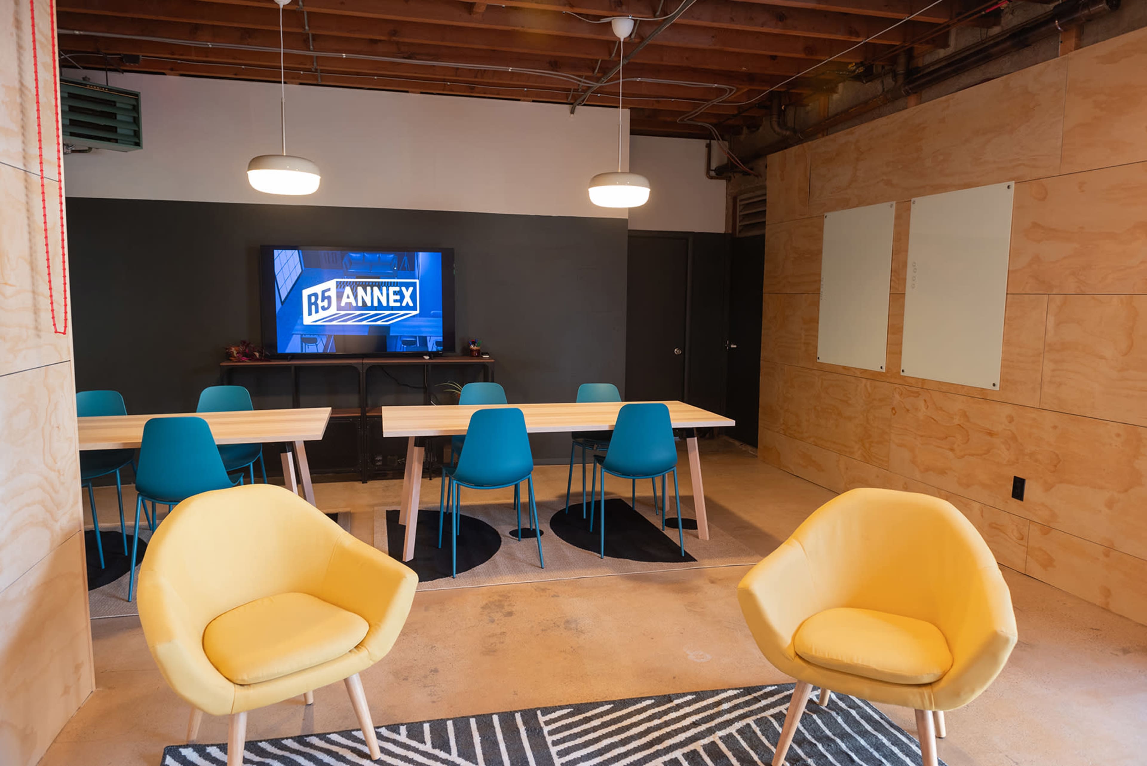 A modern meeting room with a television display, two yellow chairs, and a long table surrounded by teal chairs.