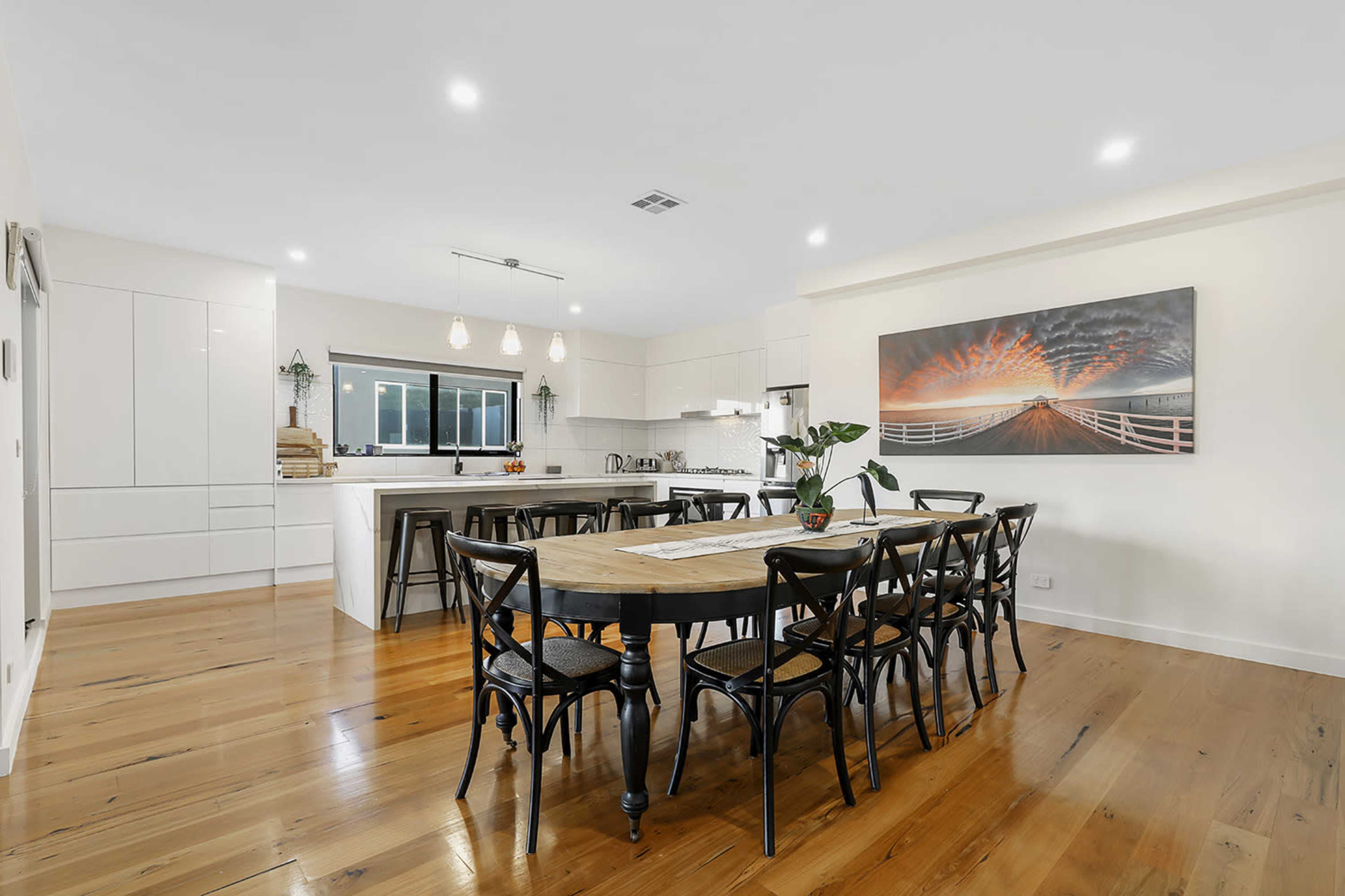 A spacious dining area features a large wooden table surrounded by black chairs, with modern cabinetry and a bright kitchen in the background.