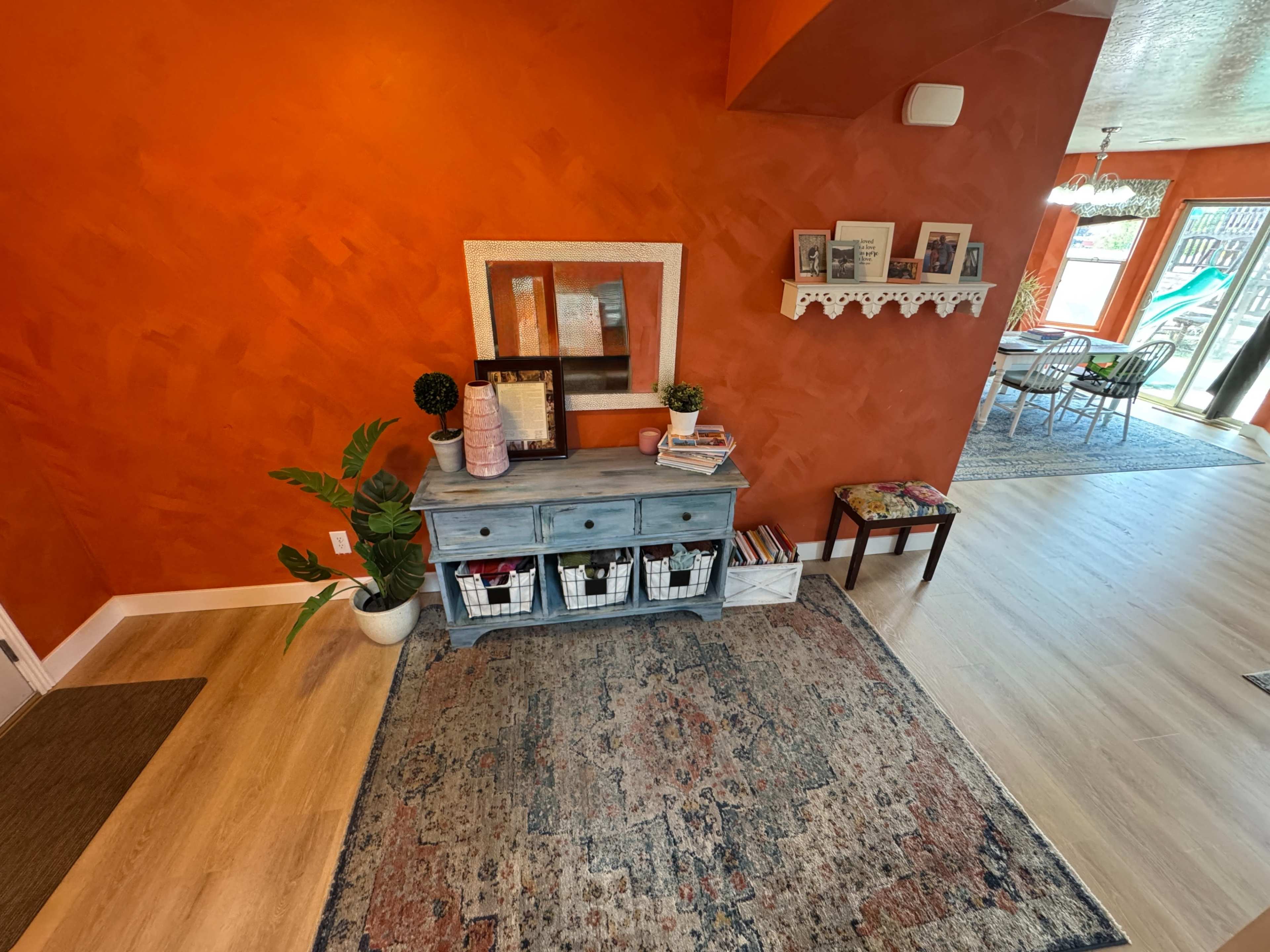 The image shows a brightly colored orange entryway featuring a blue side table with baskets, a framed mirror, and a decorative plant, along with a patterned area rug.
