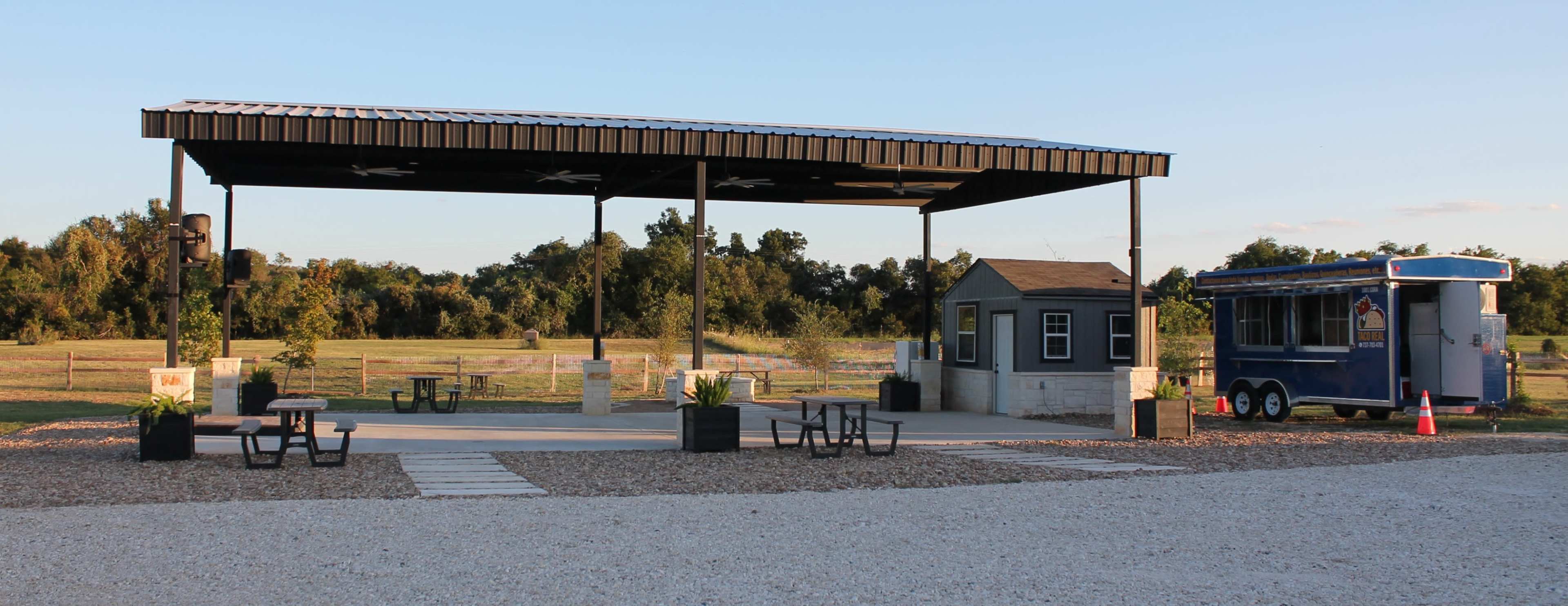 The image shows a covered outdoor seating area with tables and a food trailer parked nearby, surrounded by an open grassy landscape.
