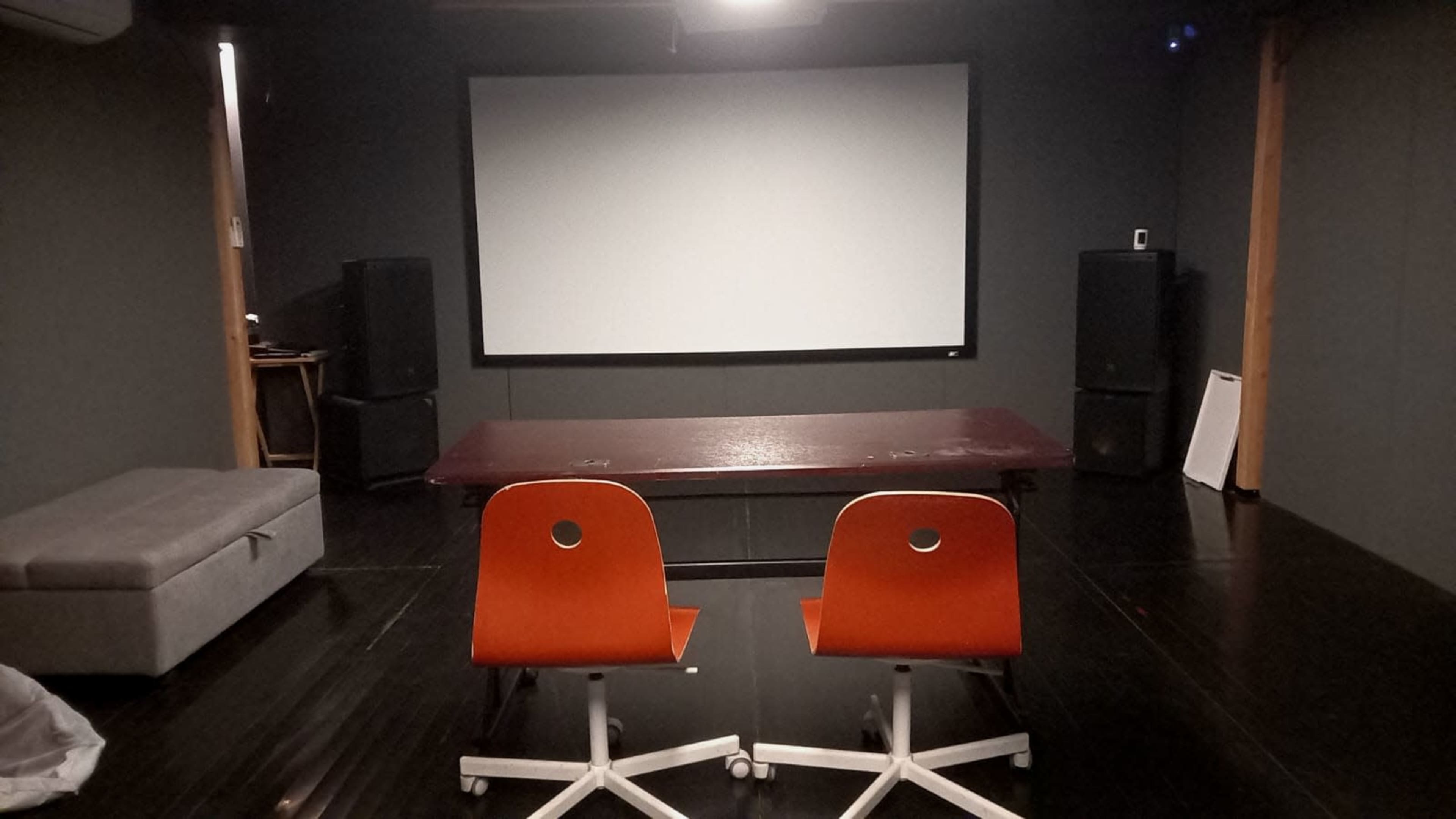 A minimalist room with a large projector screen at the front, two orange chairs facing a central table, and a gray couch in the corner.