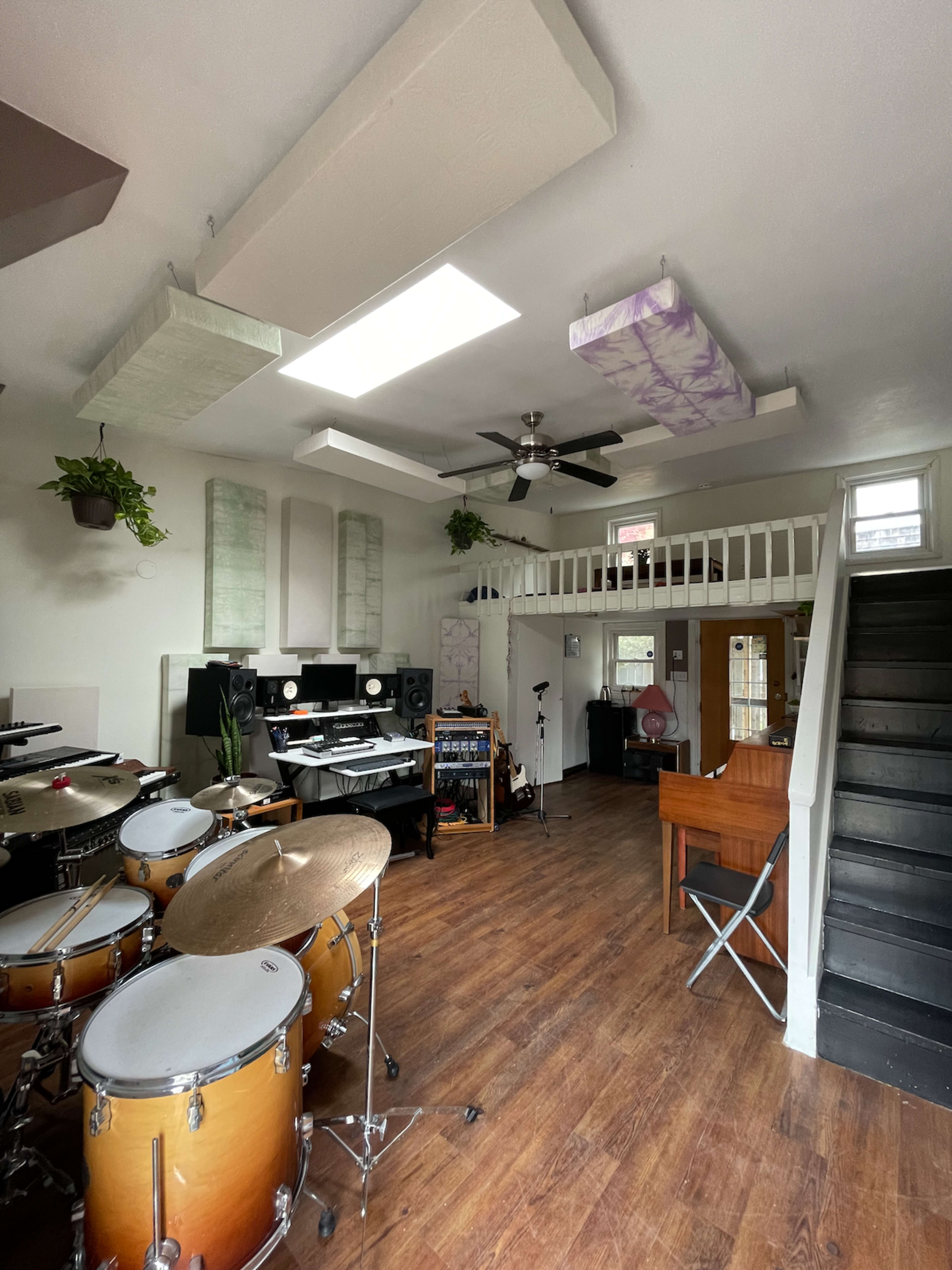 A spacious music studio featuring a drum set, a computer workstation, and a staircase leading to an upper level, with various soundproofing panels and plants decorating the space.