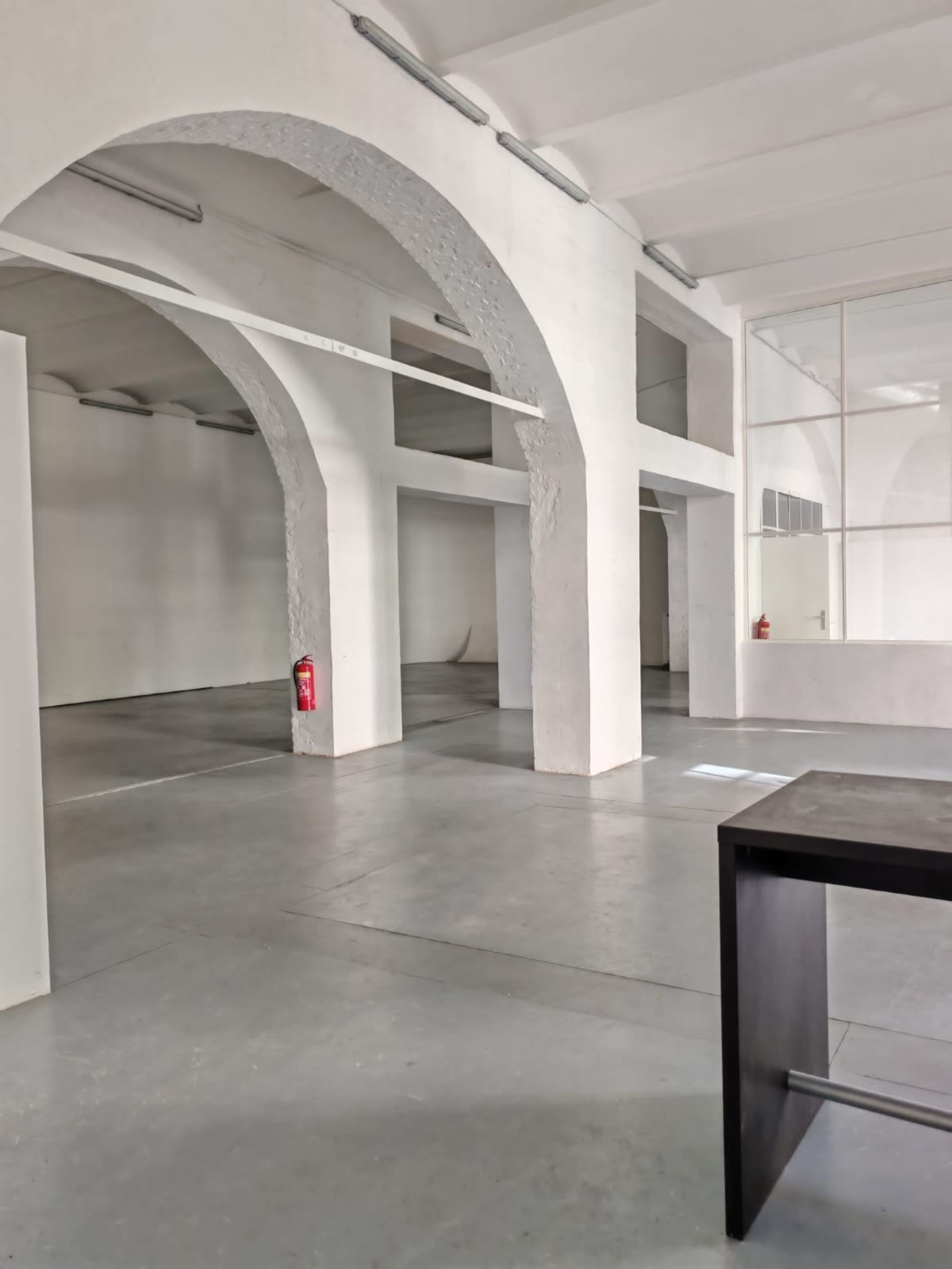 The image shows a spacious, industrial-style interior with stone arches, large open areas, and a simple black table in the foreground.