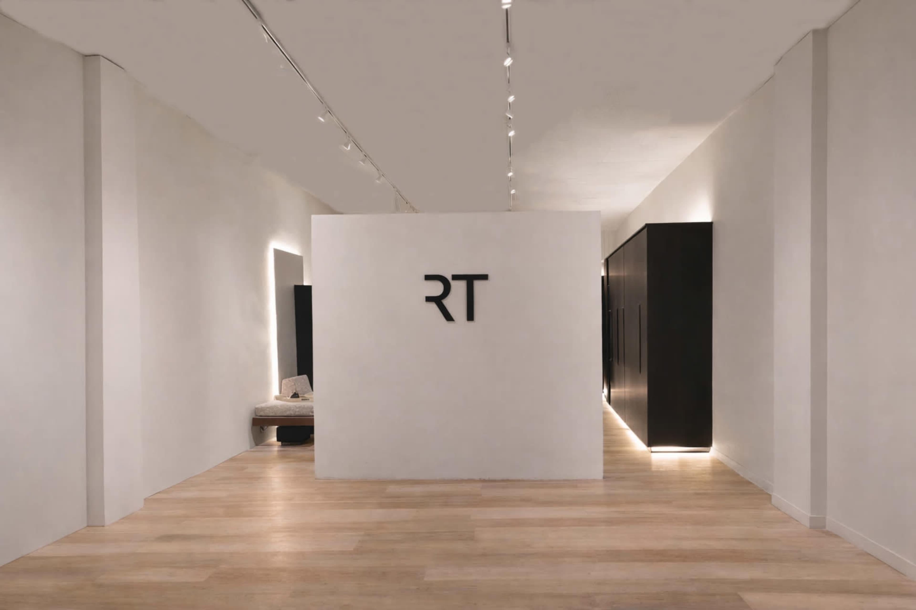The image shows a minimalistic interior space with a large white wall featuring the initials "RT," and sleek black cabinets lining the sides.
