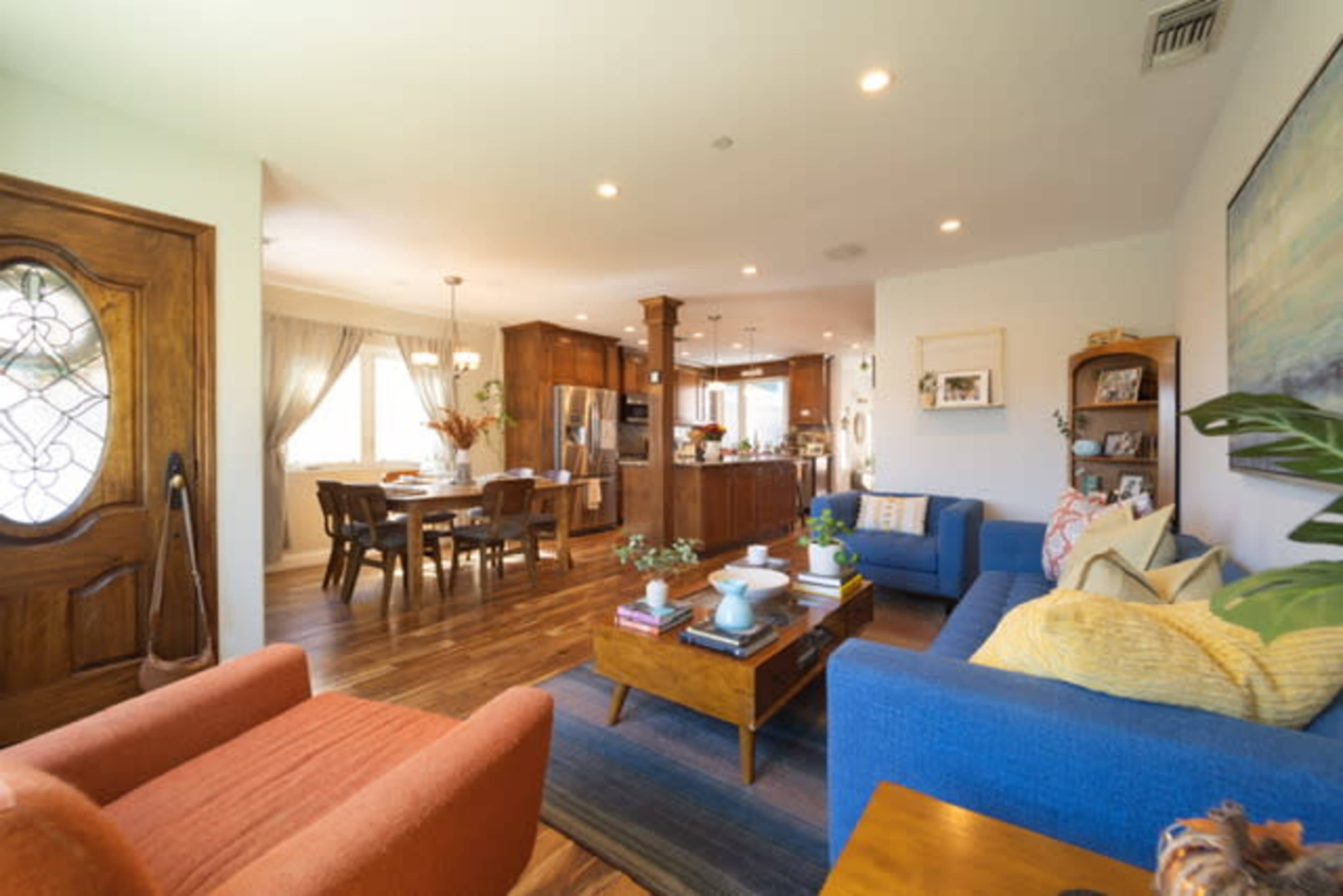 A living area with a blue couch, an orange chair, a wooden coffee table, and an open kitchen and dining area in the background.