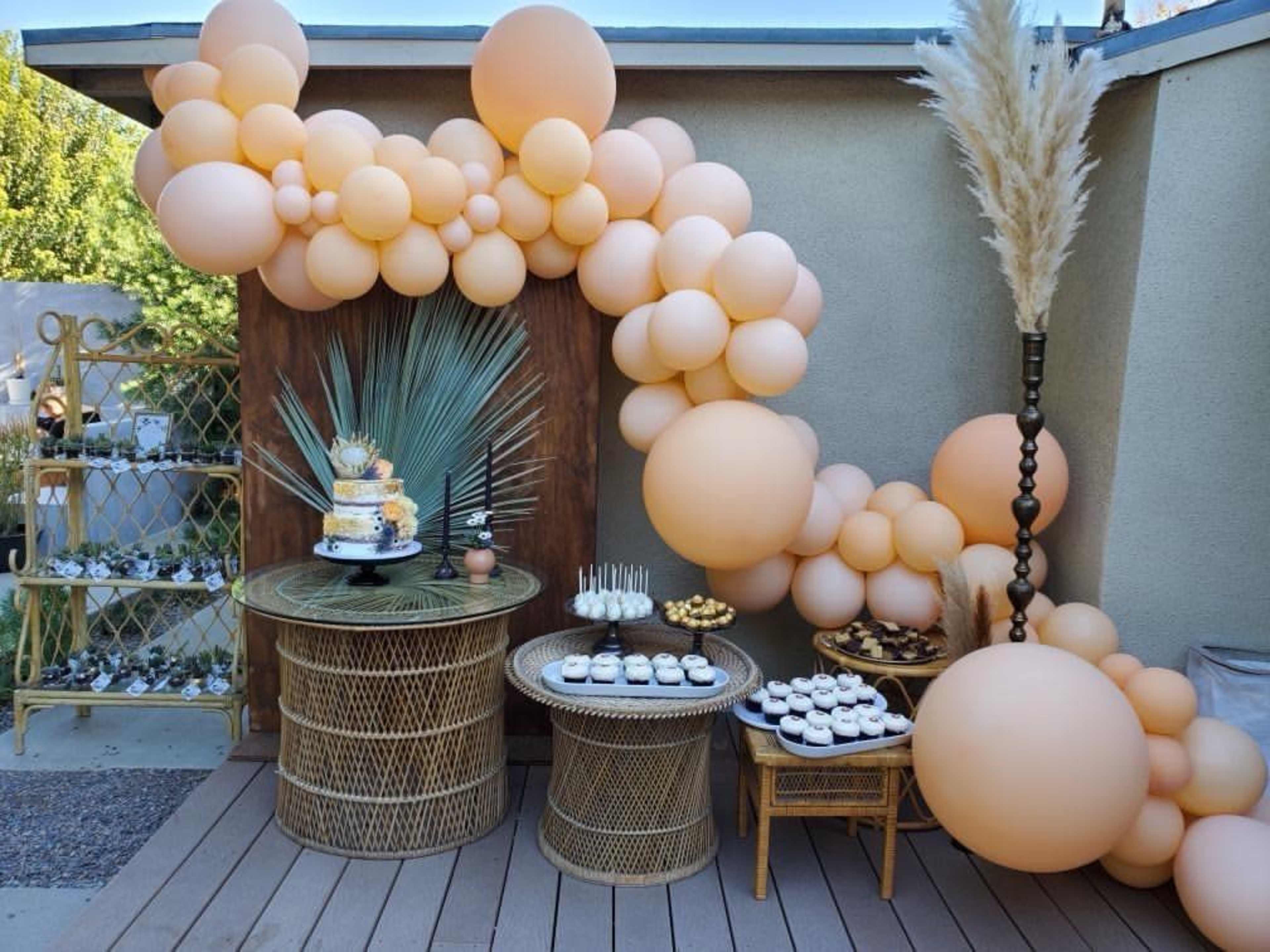 A decorated outdoor space featuring a large arrangement of peach-colored balloons, two woven tables with a tiered cake and assorted desserts.