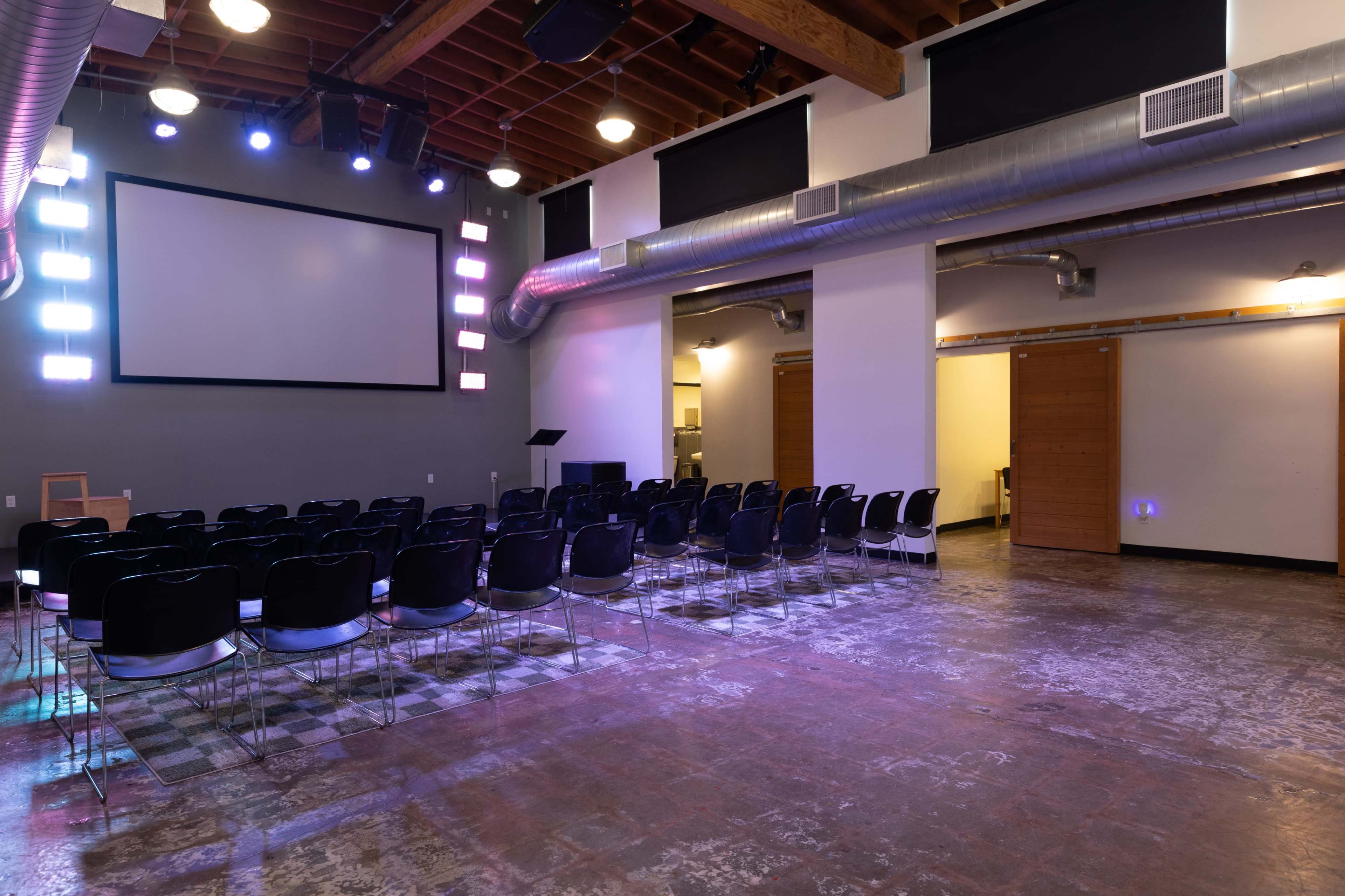 The image shows a spacious indoor venue set up with rows of black chairs facing a large screen at the front, illuminated by colorful lights and featuring exposed ductwork on the ceiling.