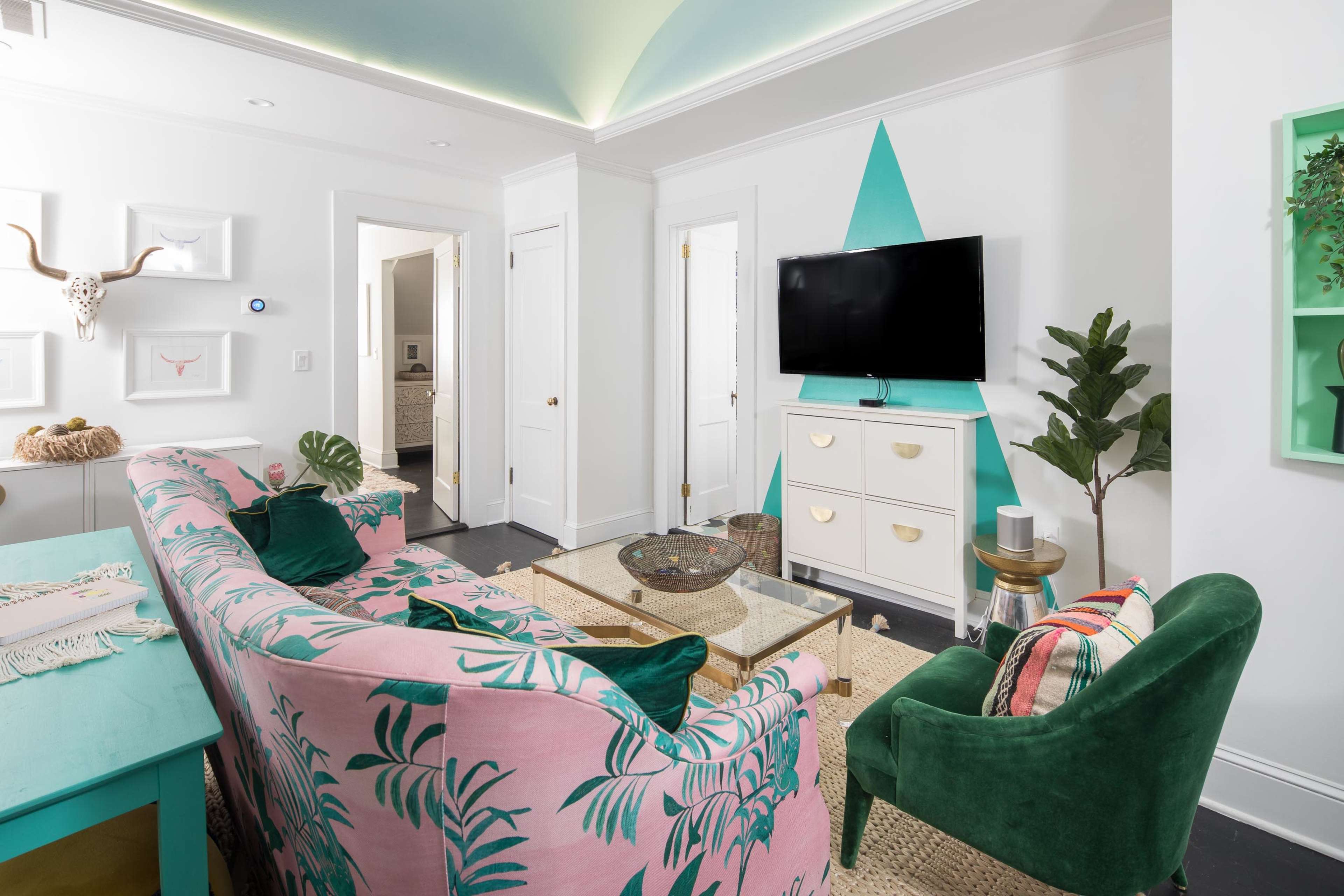 A brightly decorated living room features a pink sofa with tropical patterns, a green armchair, a glass coffee table, and a television mounted on a white wall.