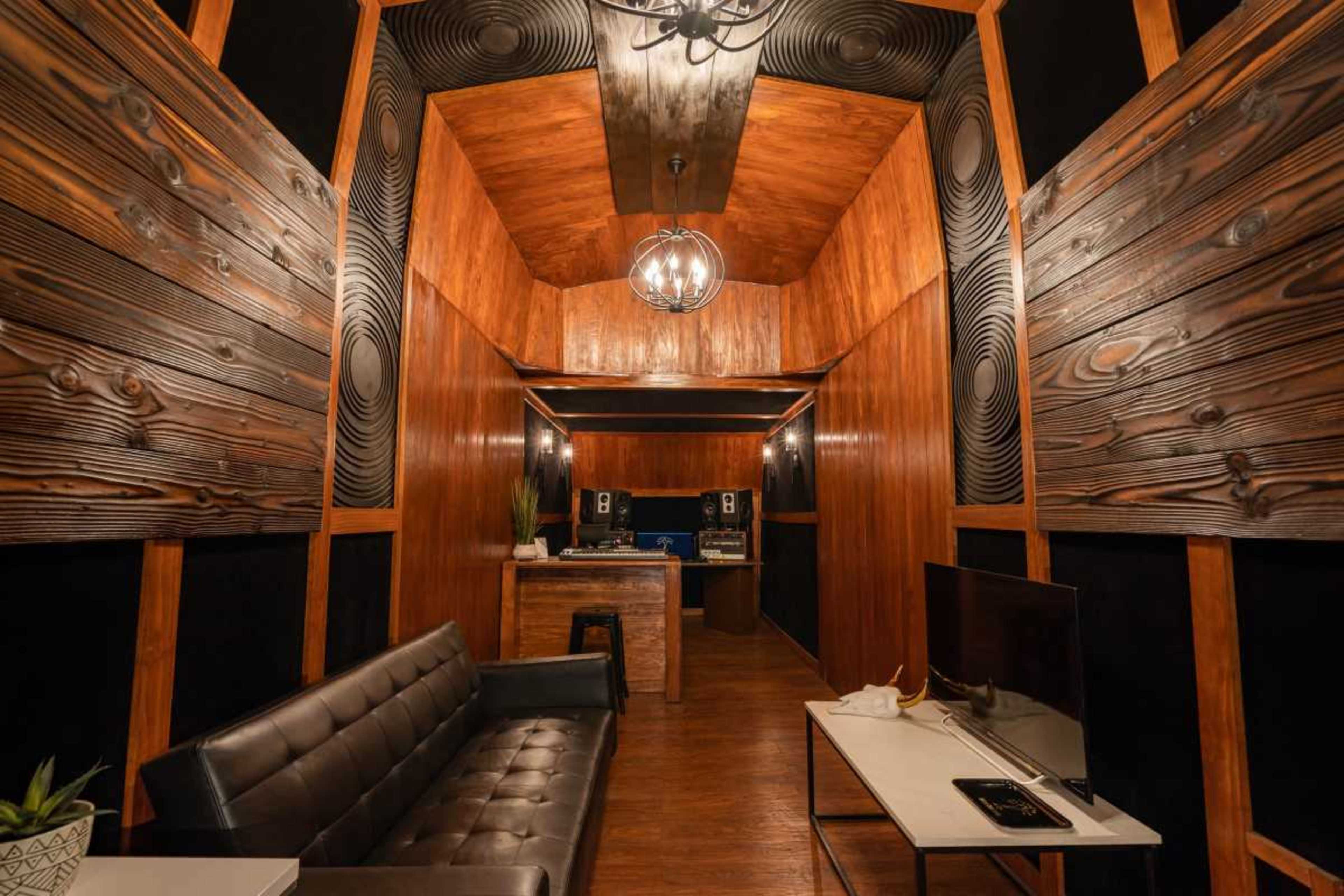 The image shows a music studio with wooden walls, a central seating area, and audio equipment visible in the background.