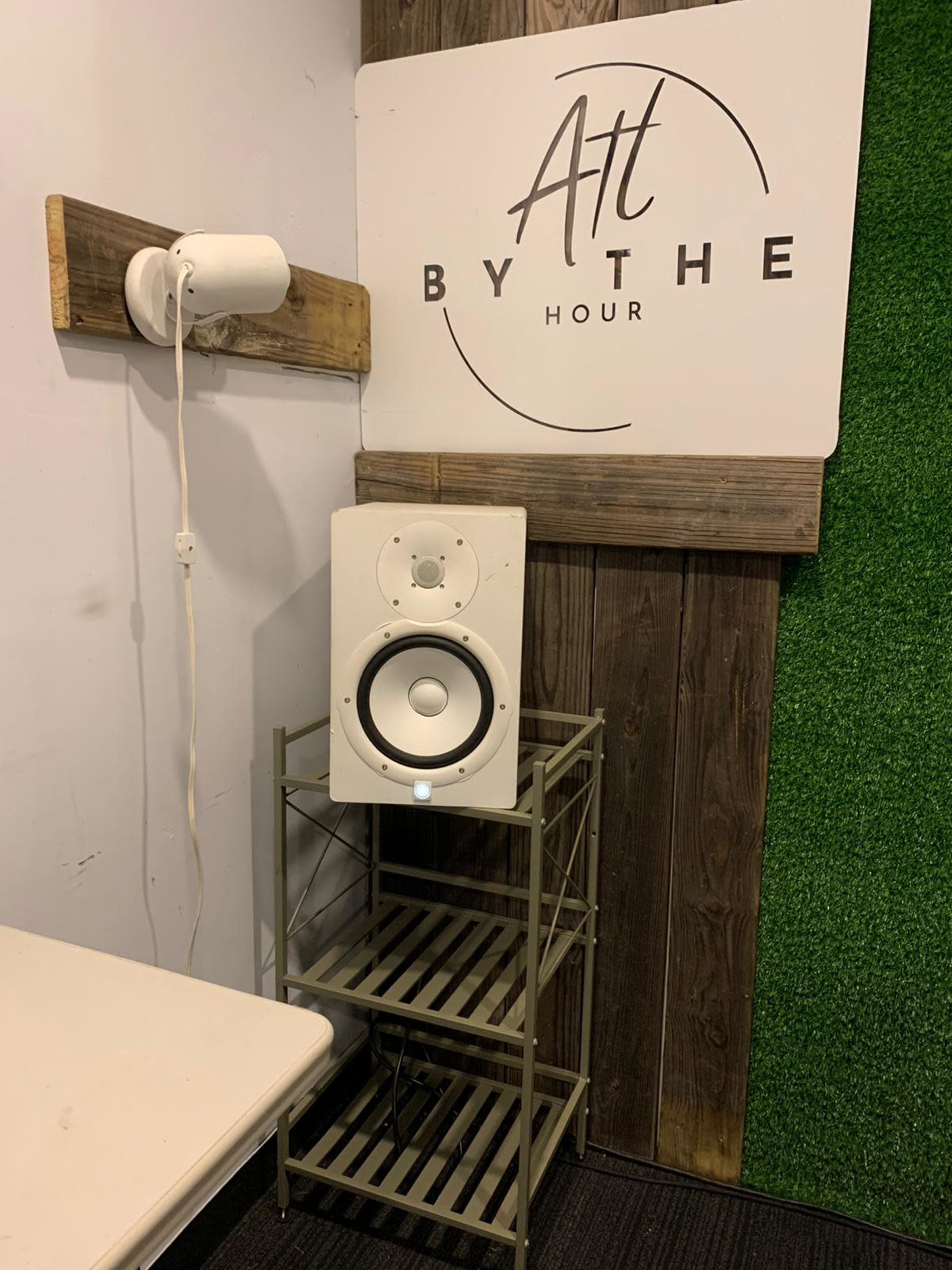 A white speaker on a metal shelving unit next to a wooden wall, beneath a sign that reads "Art By The Hour."