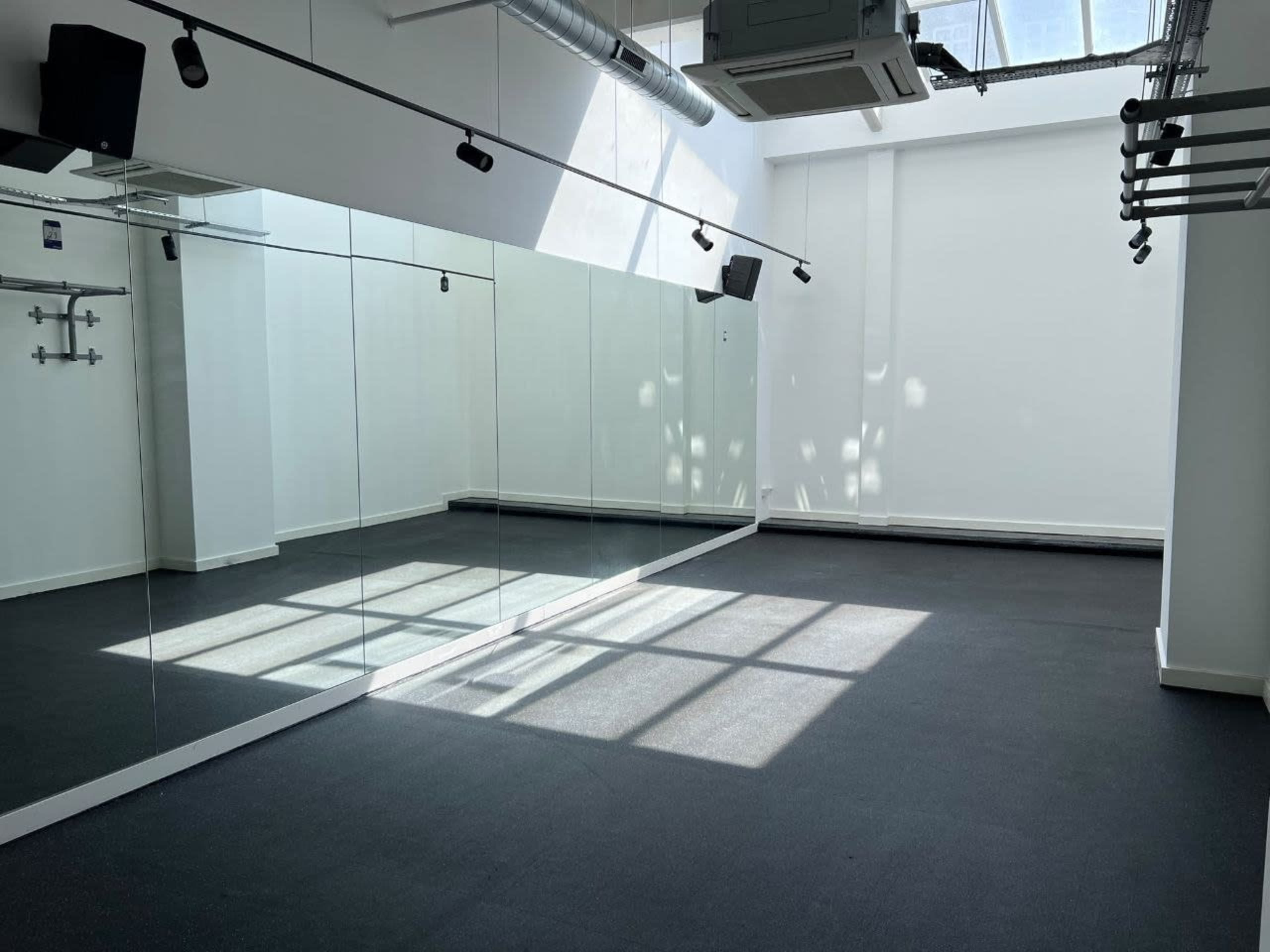 The image shows a bright, empty studio space with large mirrors on one wall and a black rubber floor.