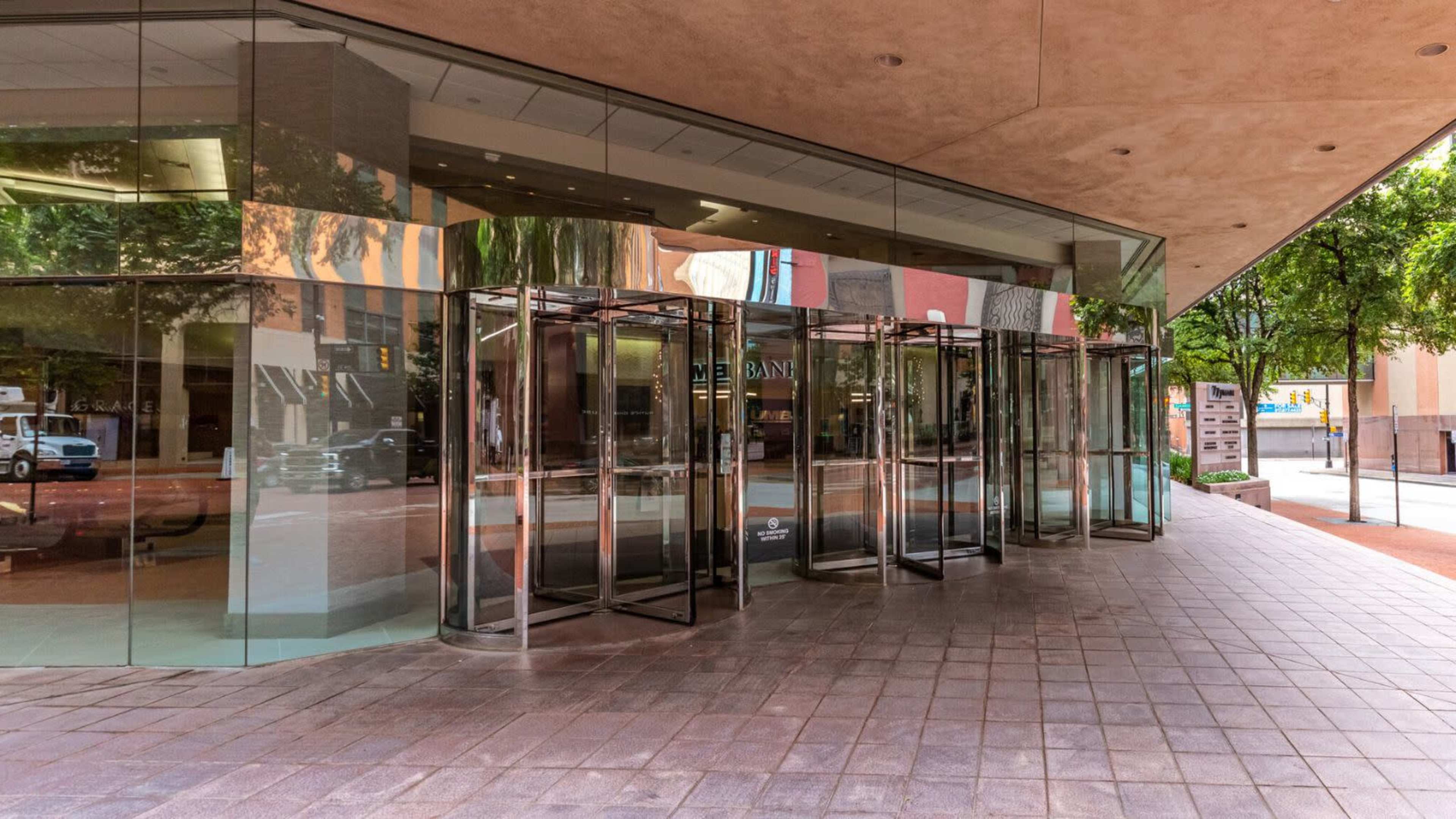 The image shows a modern glass entrance with revolving doors on a street lined with trees and buildings.