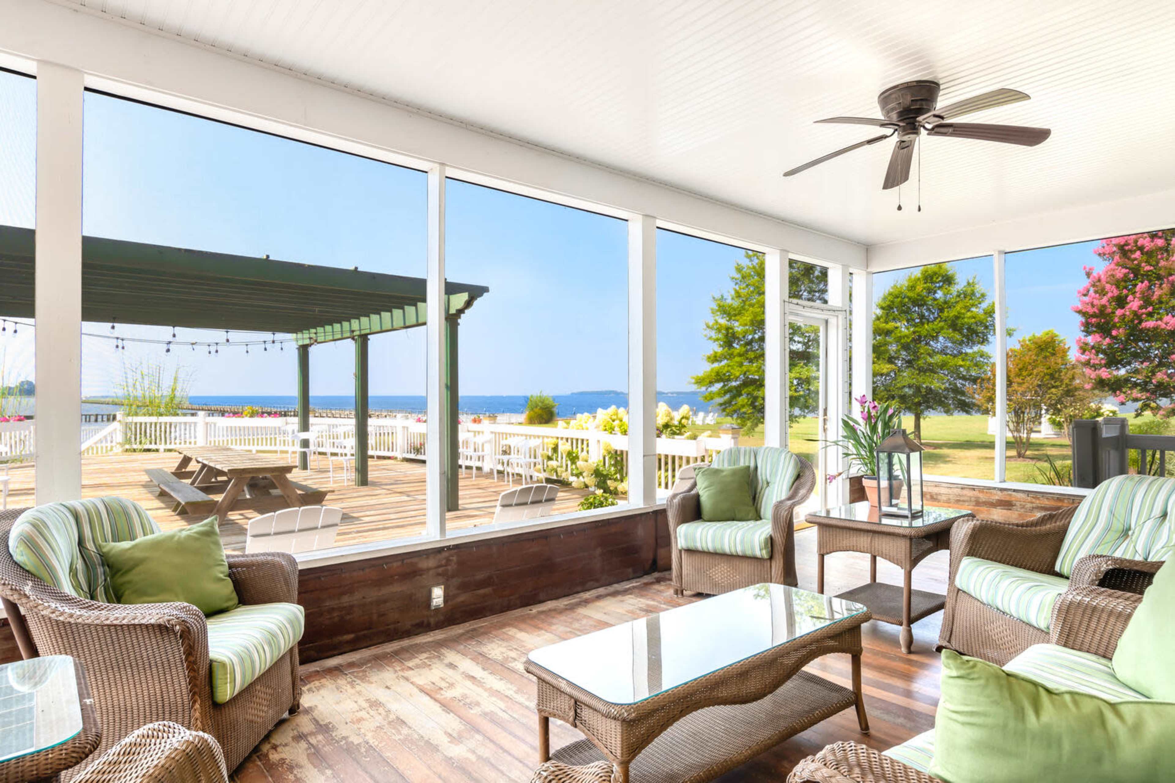 A screened-in porch features wicker furniture and a glass table, overlooking a deck and a view of a body of water and trees.