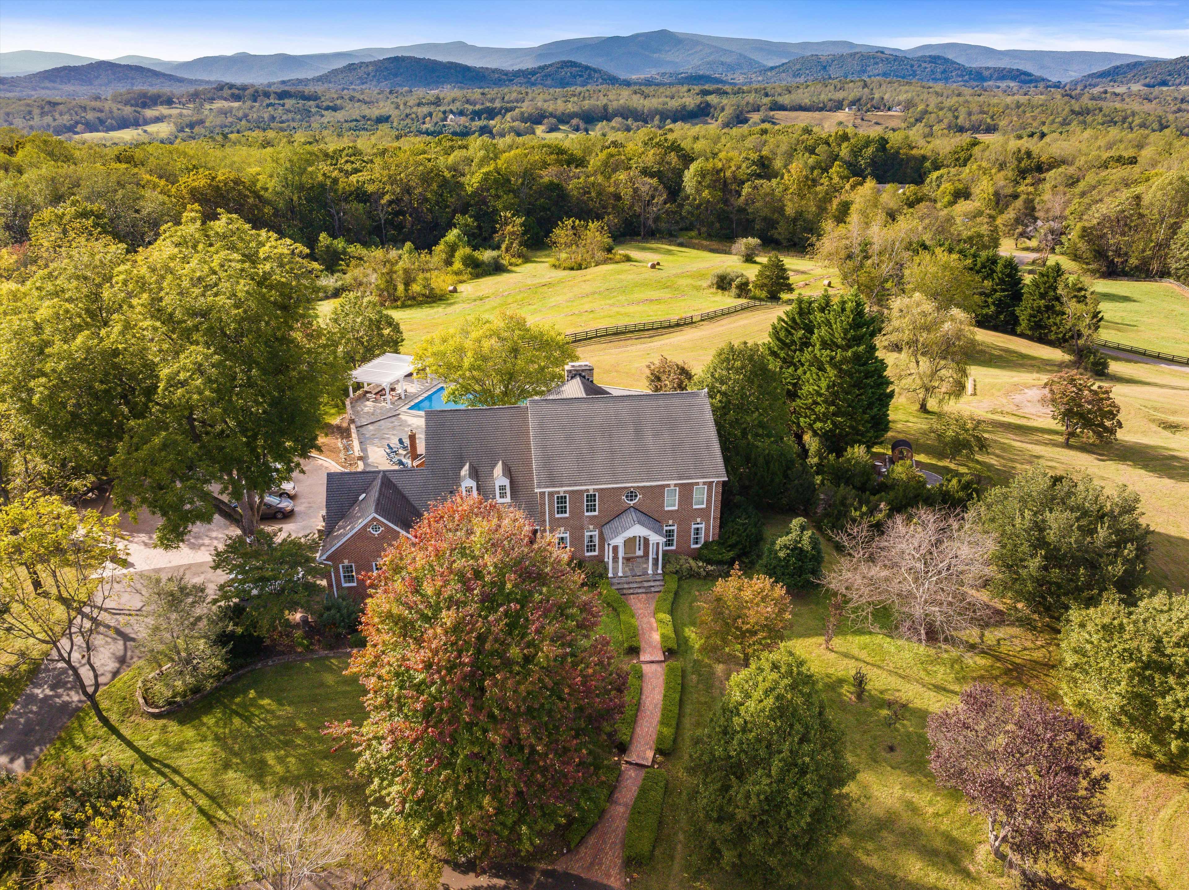 The image shows a large, elegant house surrounded by green fields and trees, with mountains in the background.