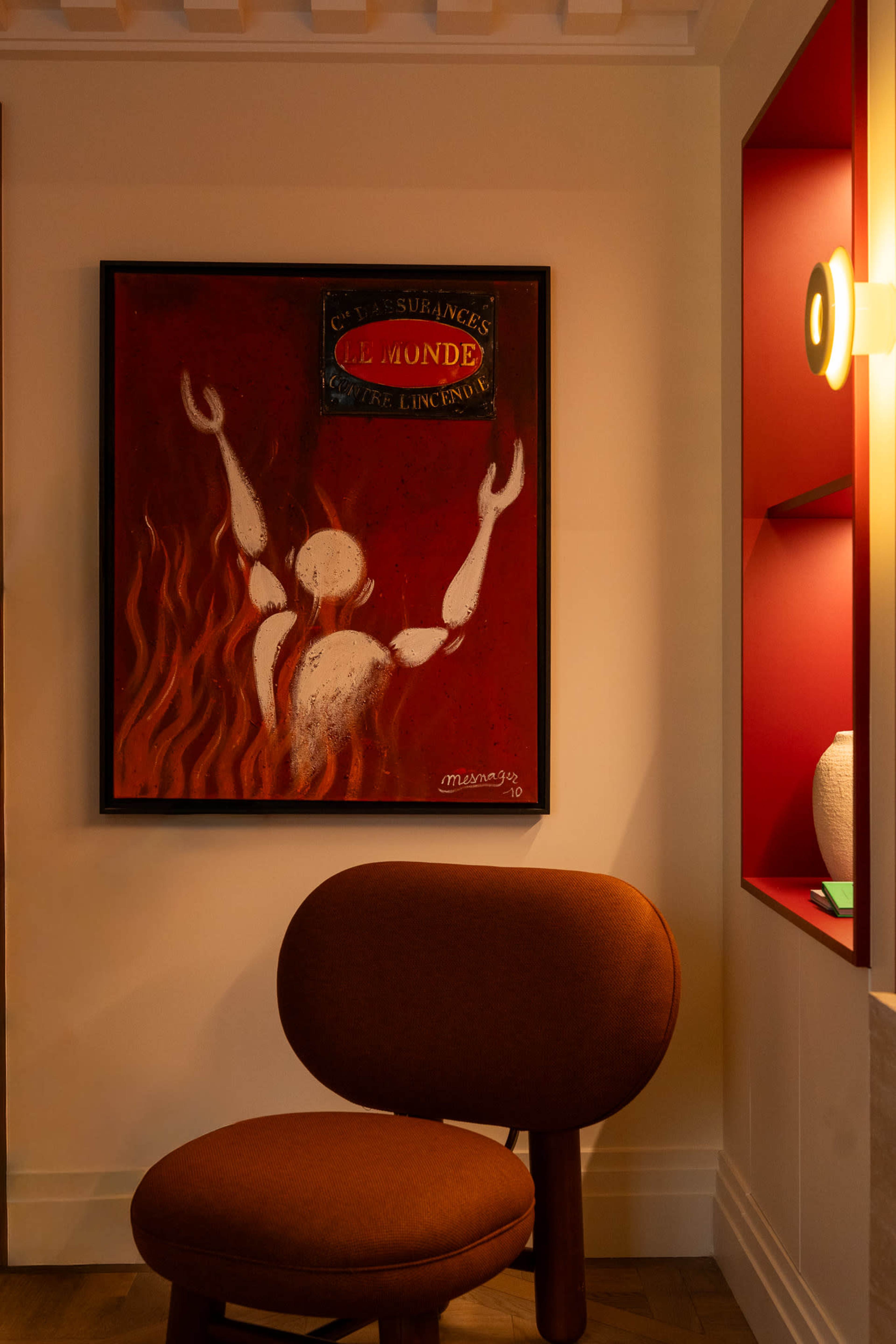 The image shows a framed artwork featuring a red and white figure with raised arms on a wall beside a brown circular chair and a lit wall sconce.