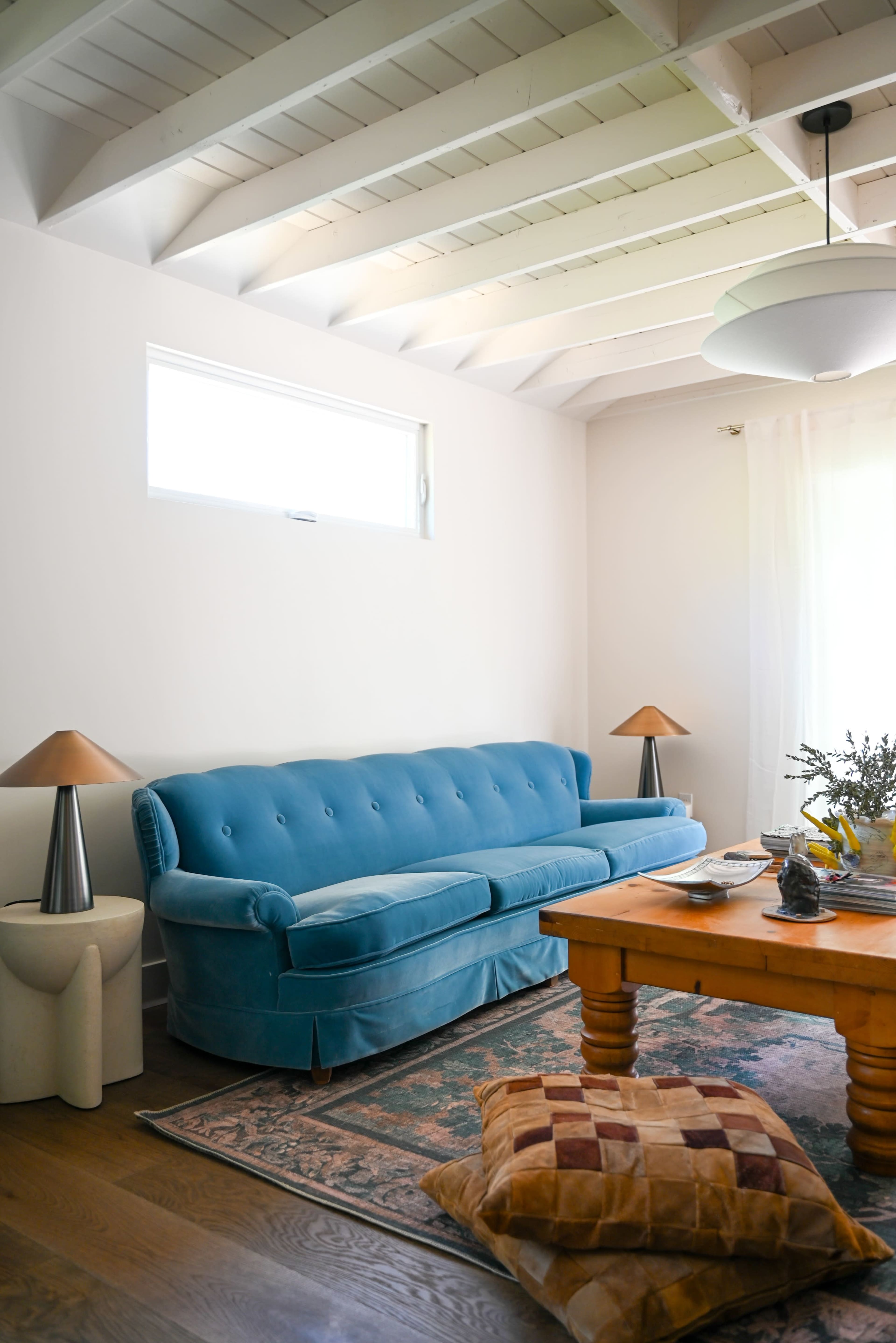 A bright living room features a blue velvet sofa, a wooden coffee table, two table lamps, and a large window allowing natural light.