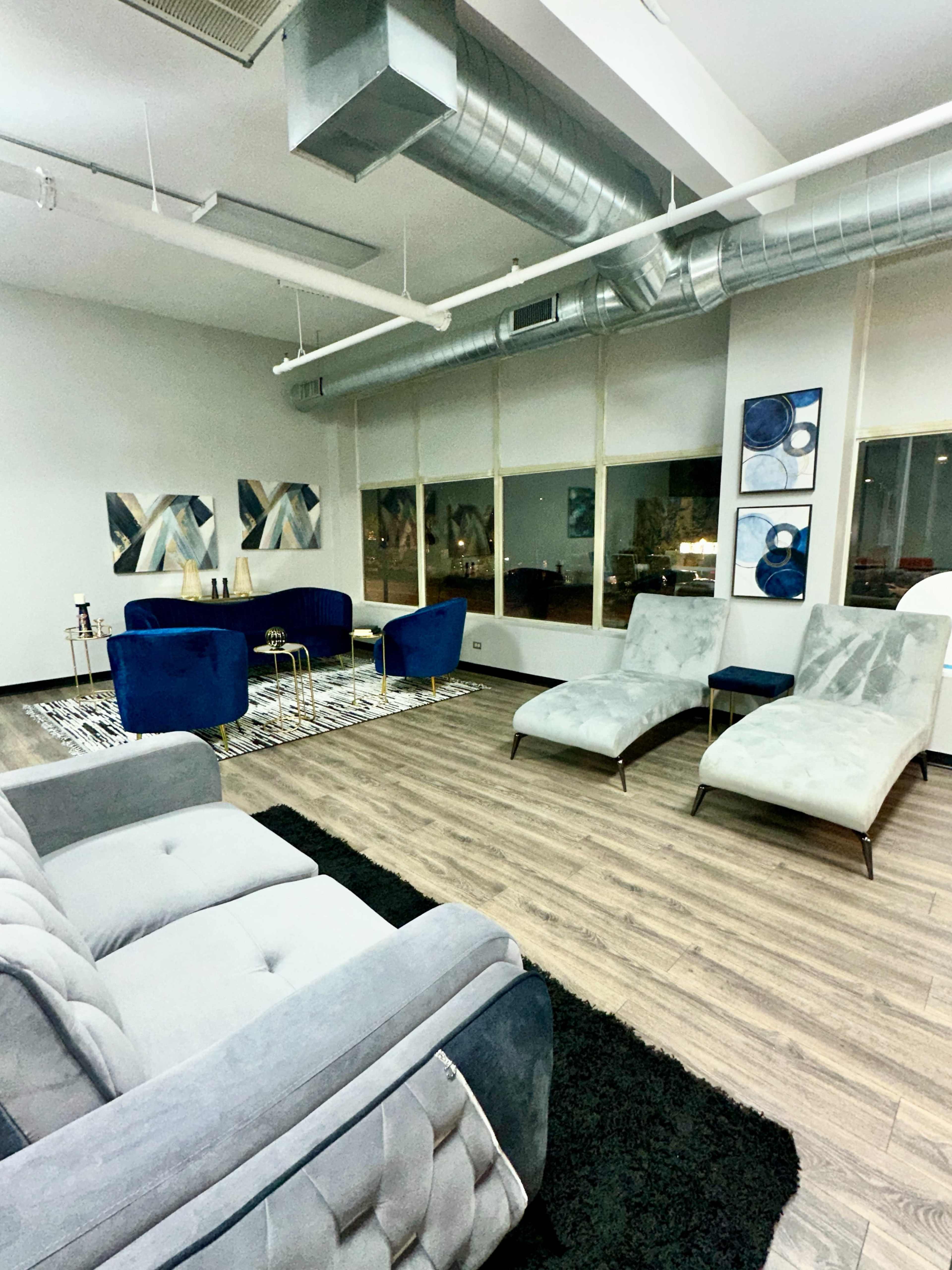 The image shows a modern lounge area with gray and blue furniture, including sofas and chaise lounges, set against a backdrop of large windows.