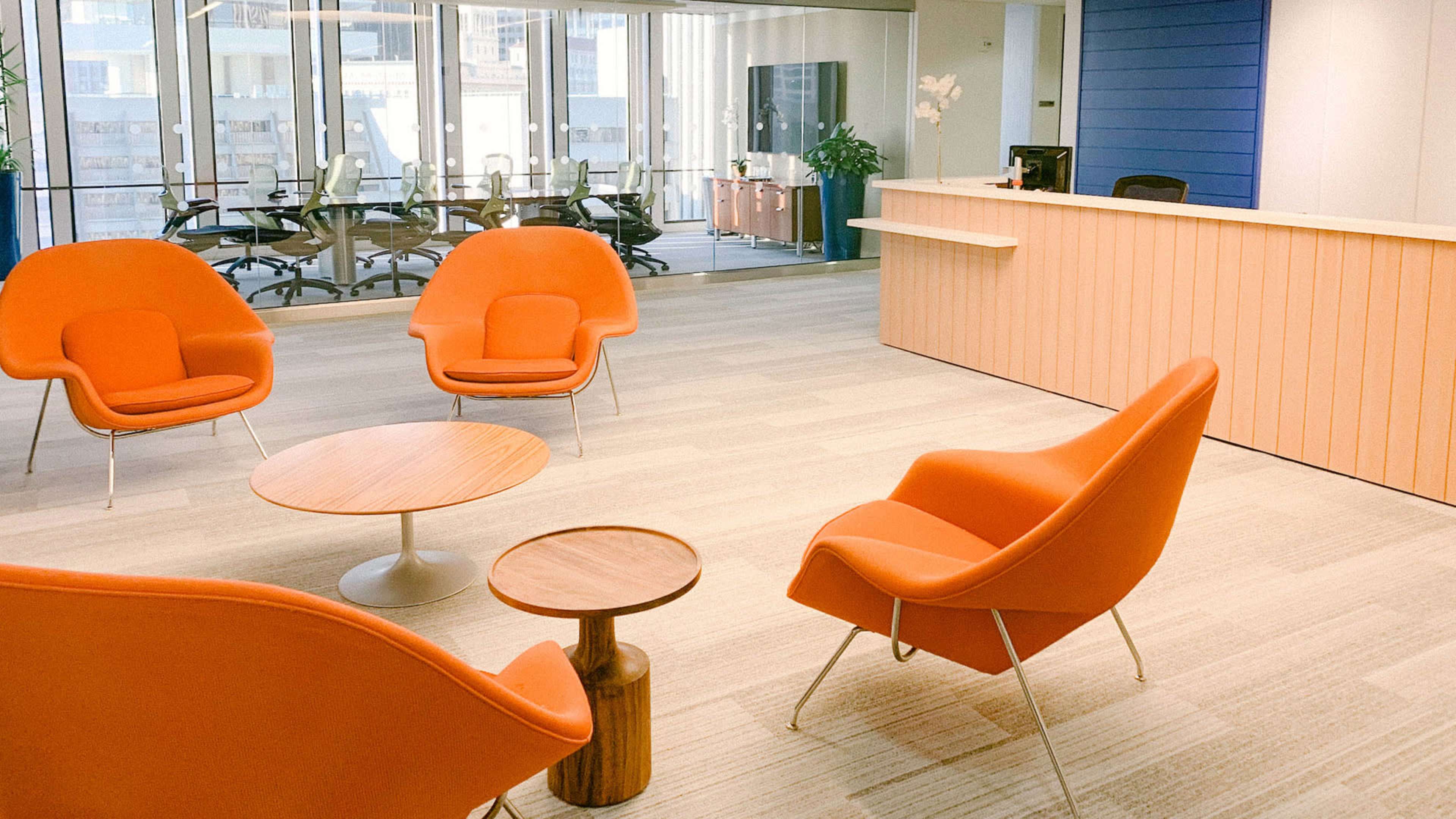 A modern office reception area with orange chairs and a wooden coffee table, featuring a large glass window that overlooks a conference room.
