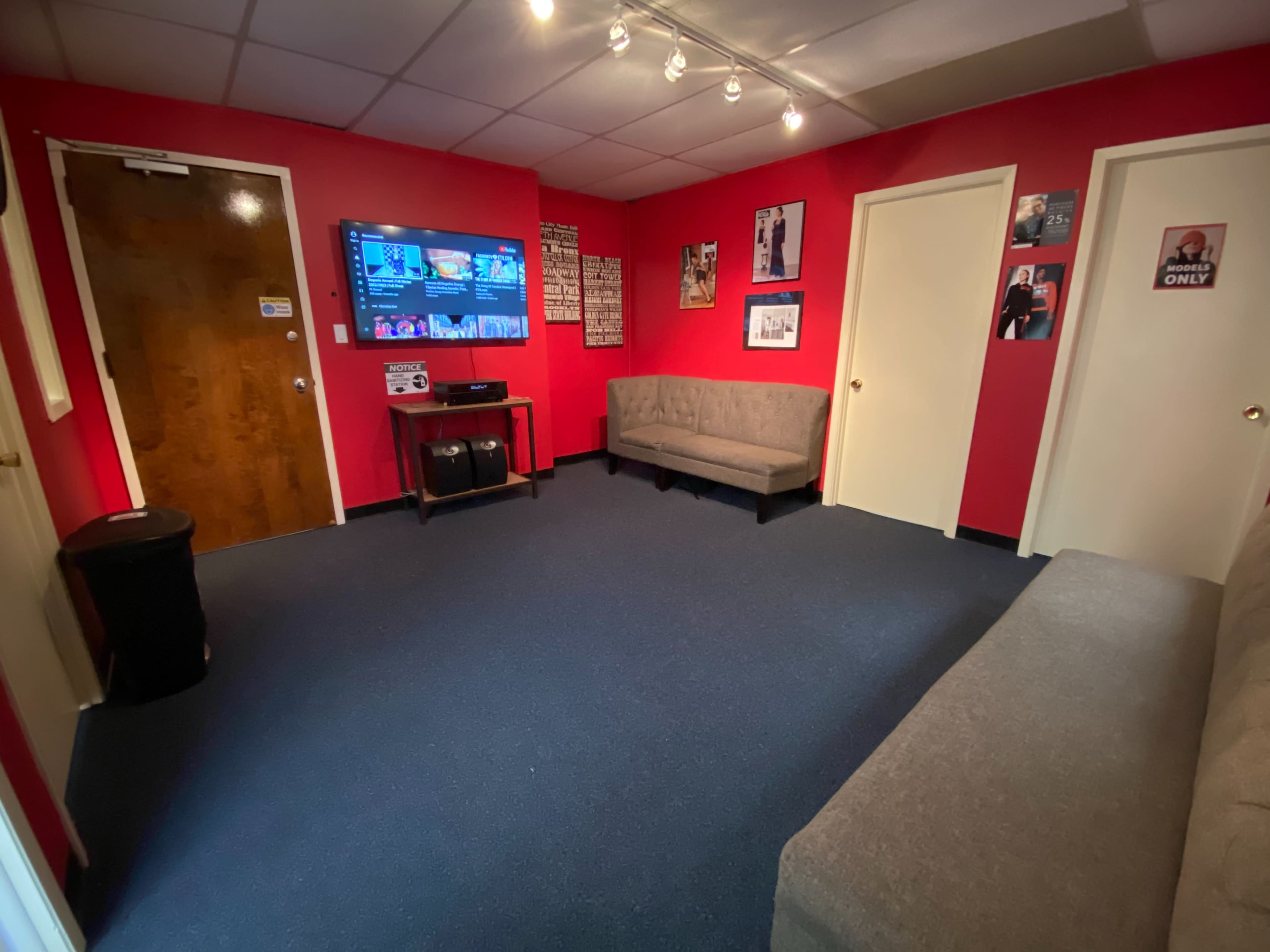 The image shows a small waiting room with red walls, two sofas, a television mounted on the wall, and a door leading outside.