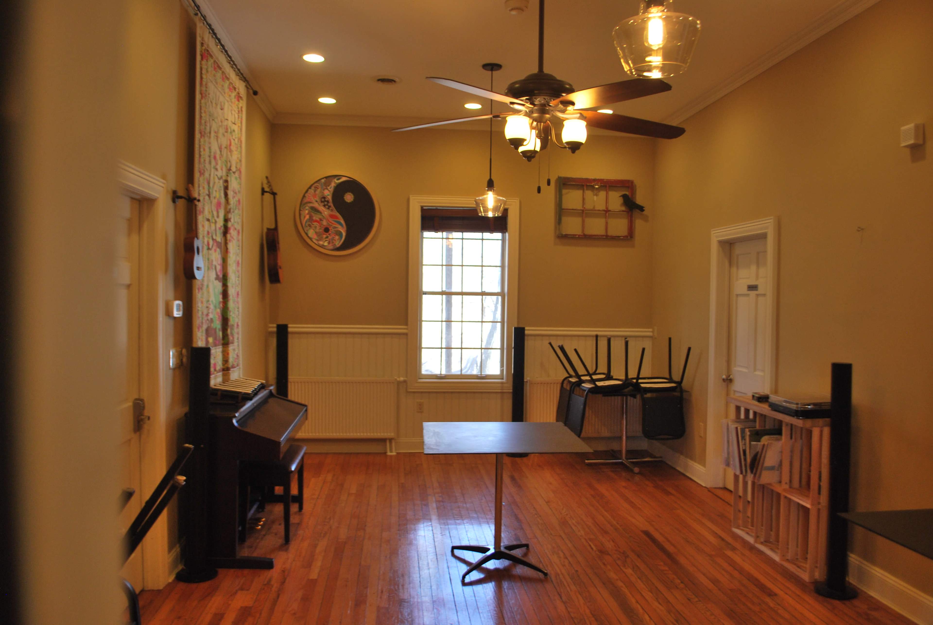A spacious room with wooden floors, a ceiling fan, a table in the center, and some furniture stacked against the walls.
