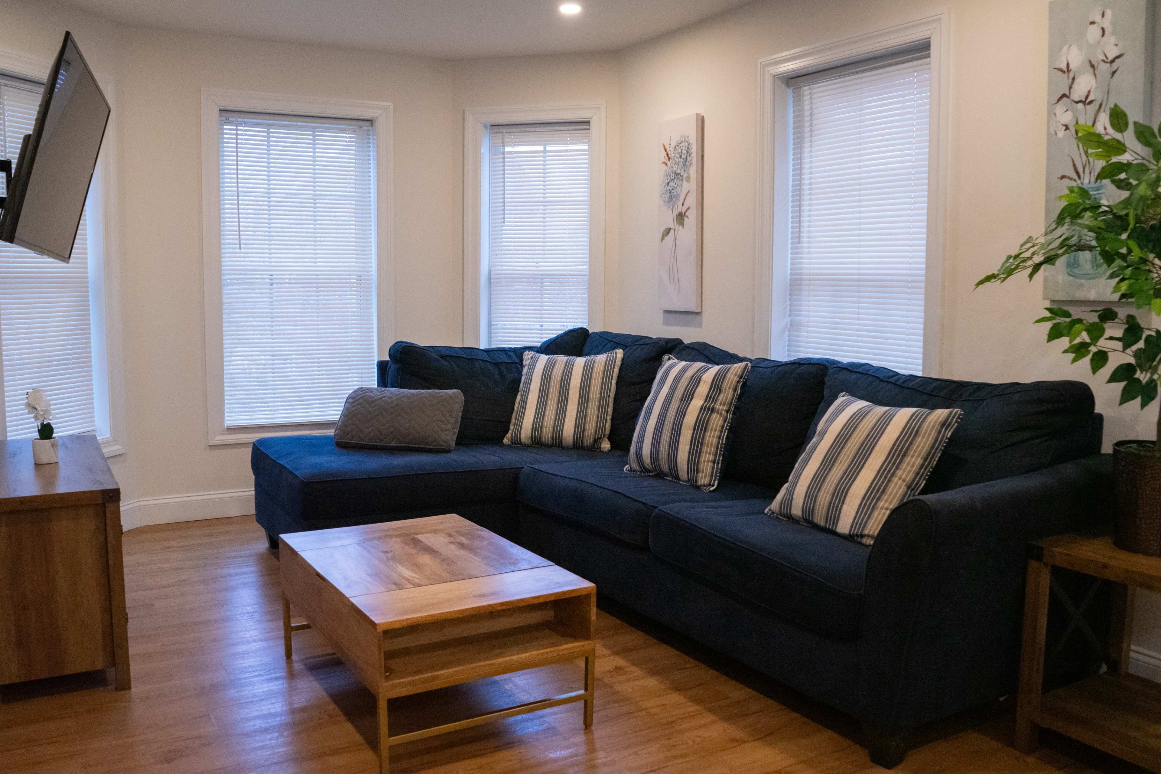 A living room featuring a dark blue sectional sofa with striped cushions, a wooden coffee table, and large windows with blinds.