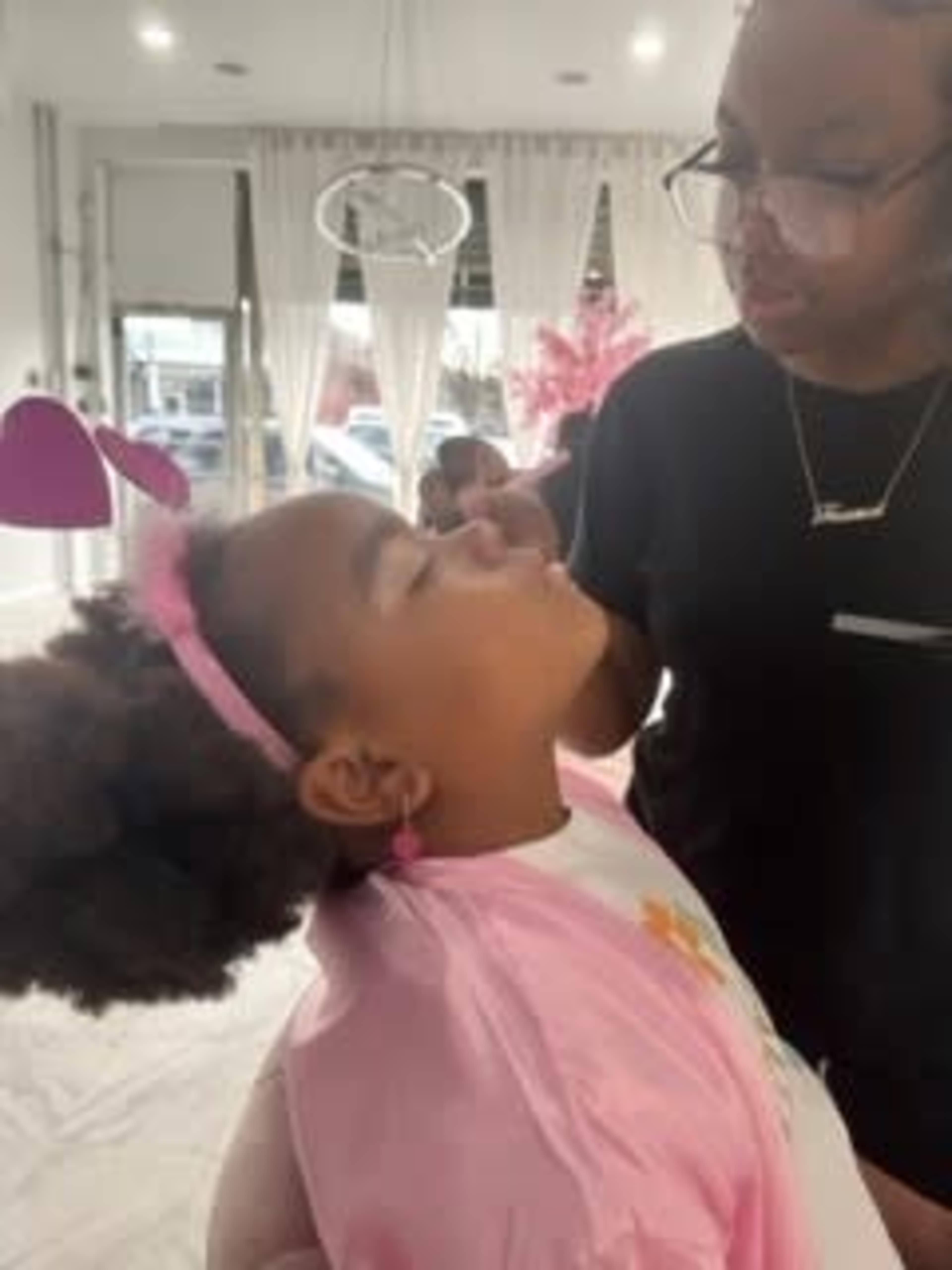 A young girl wearing a pink headband is having her makeup applied by an adult in a bright, decorated room.