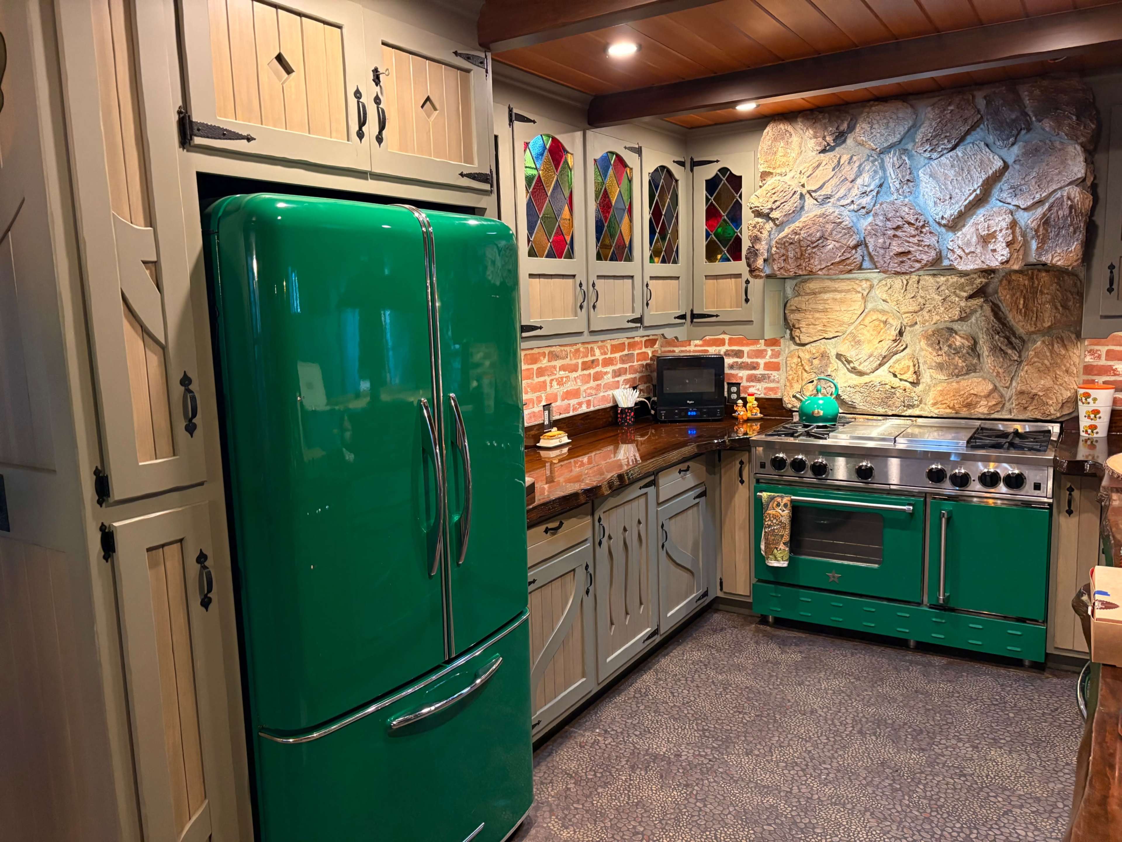 The image shows a kitchen featuring vintage-style green appliances, including a refrigerator and a stove, set against a backdrop of stone and brick walls.