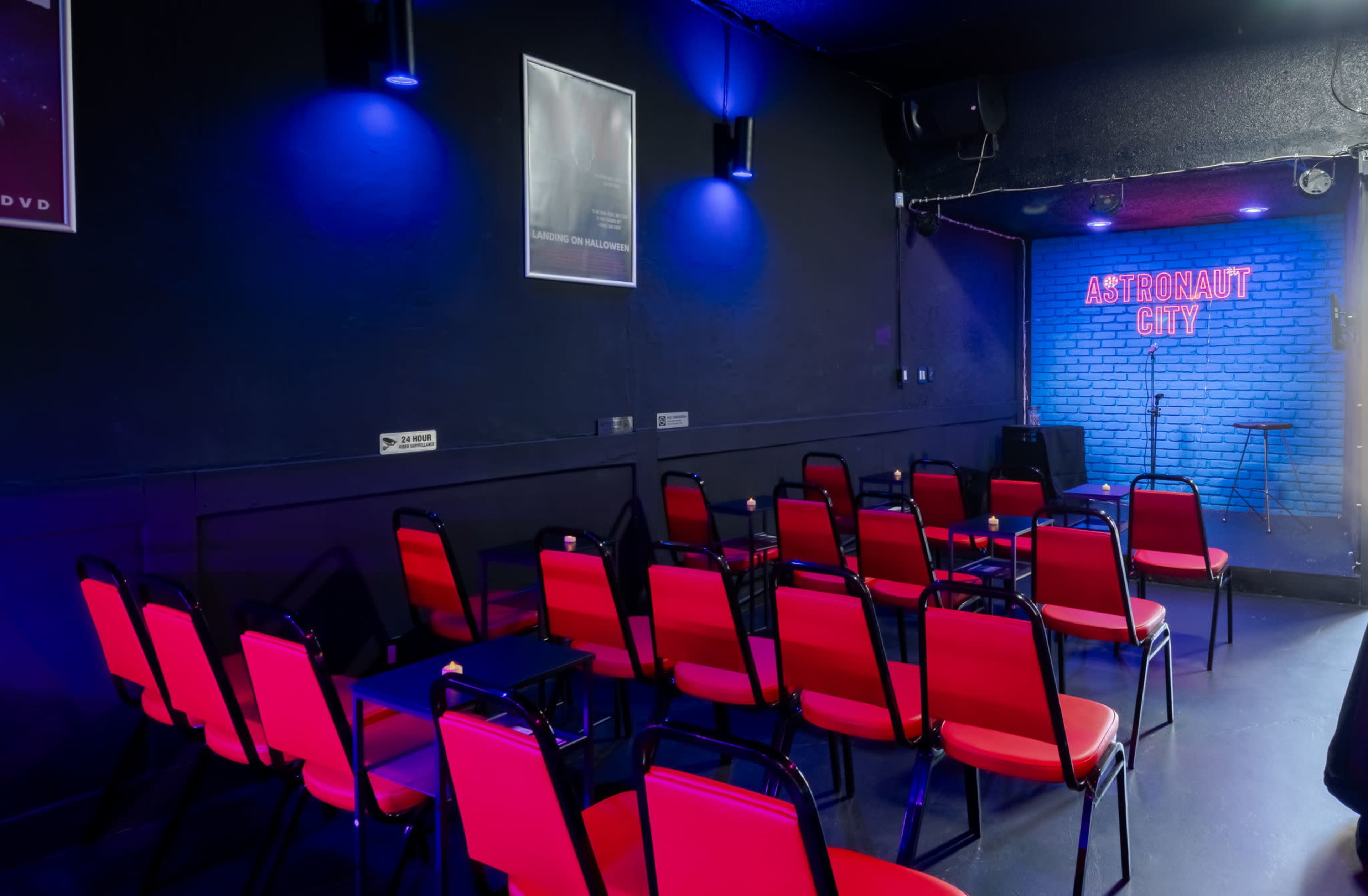The image shows an empty comedy club with red chairs arranged in rows facing a blue-brick stage that has a microphone stand.