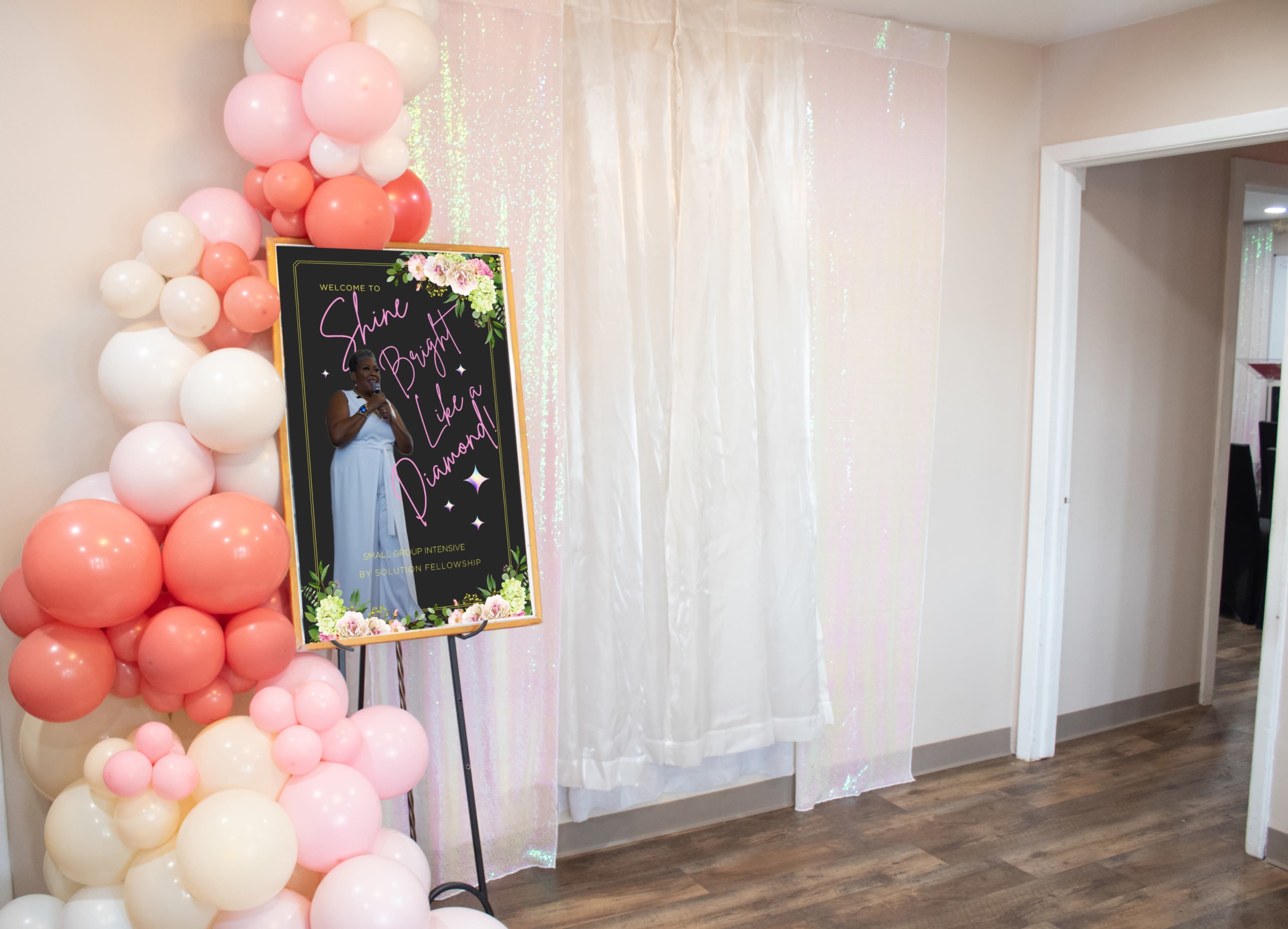 The image features a decorative setup with a balloon arch in shades of pink and cream beside a welcome sign against a shimmering backdrop.