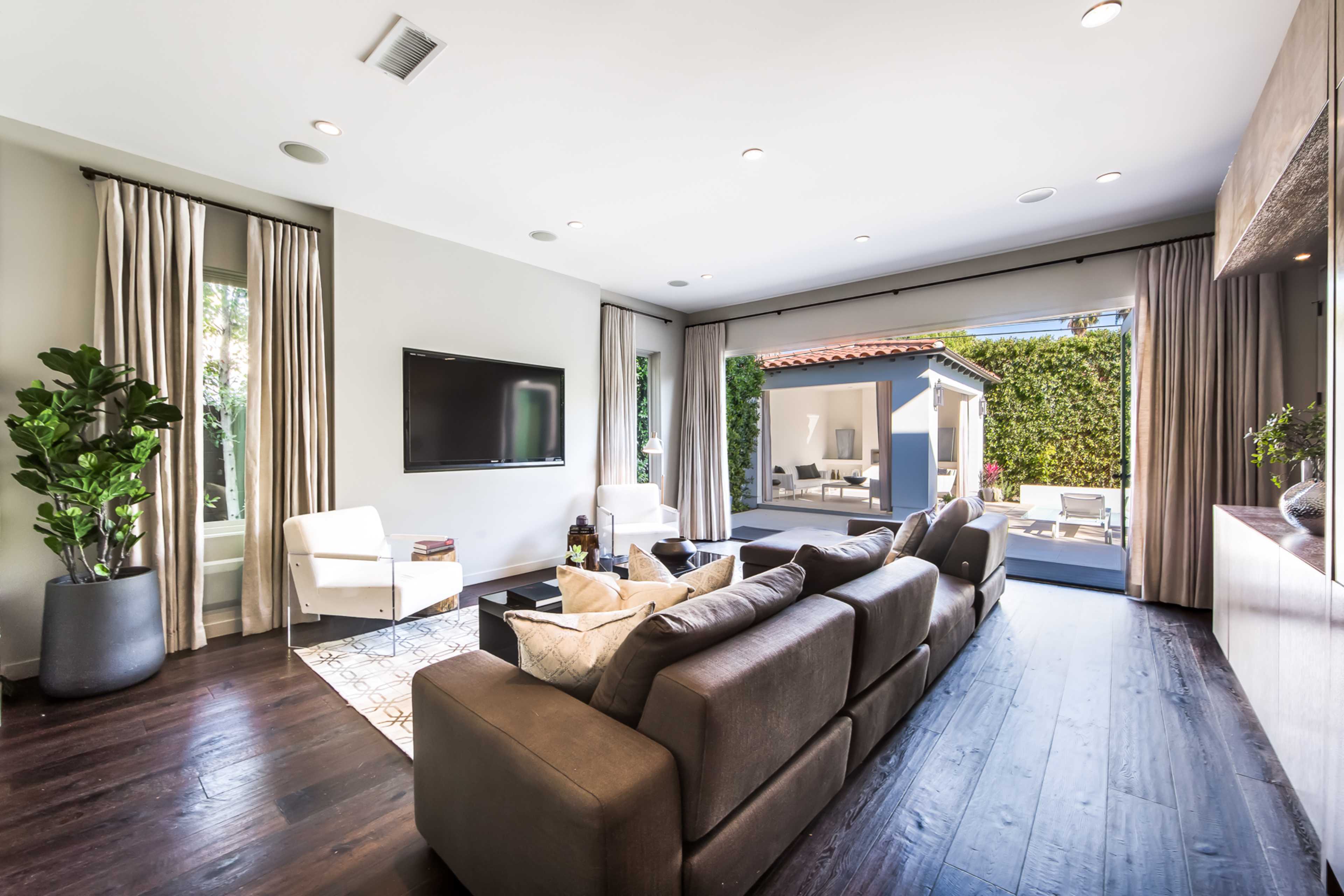 The image shows a modern living room with a dark leather sofa, a large television mounted on the wall, and sliding glass doors leading to an outdoor area.