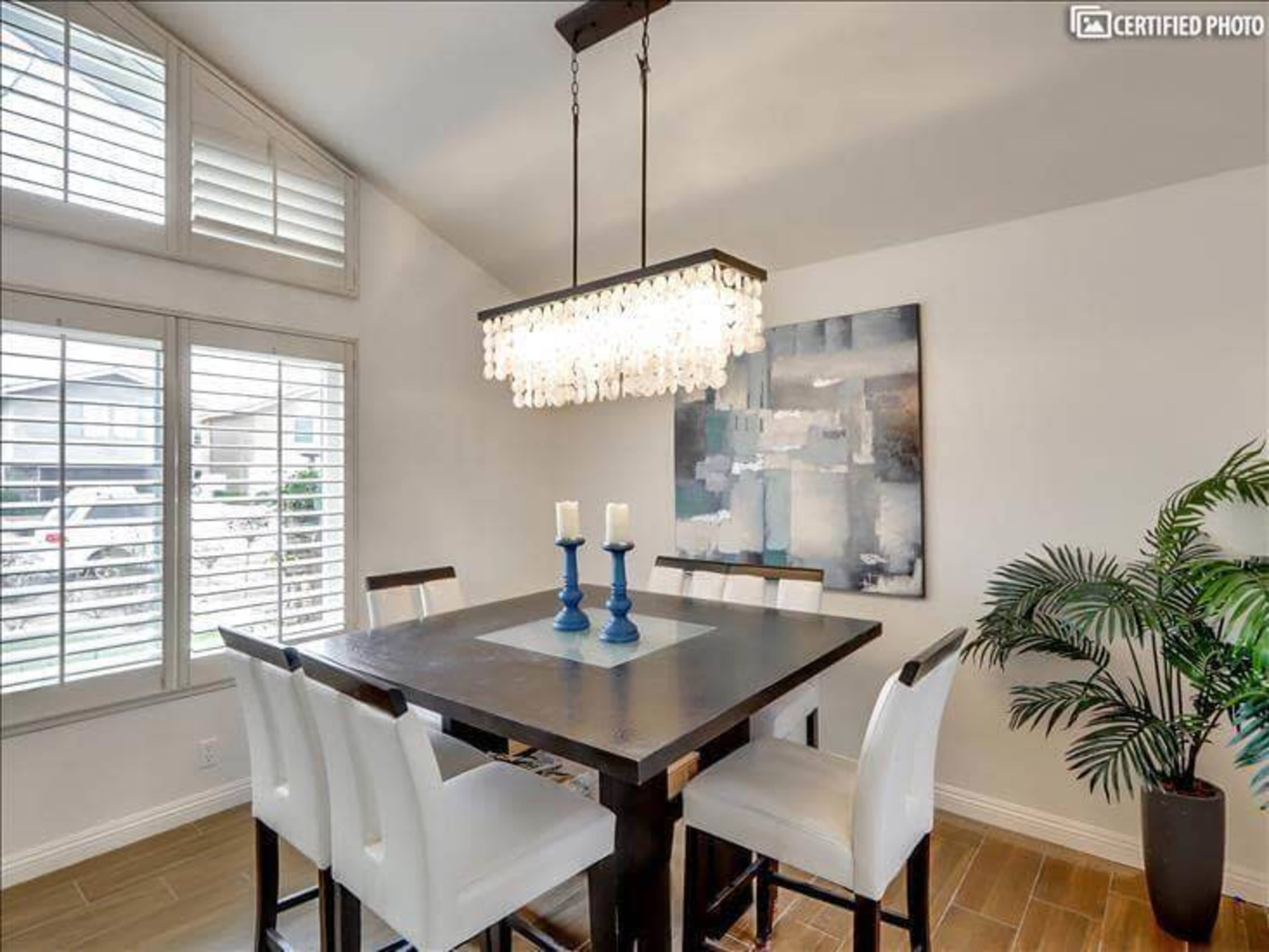 A dining area features a dark wooden table surrounded by white chairs, with a modern chandelier above and a large artwork on the wall.