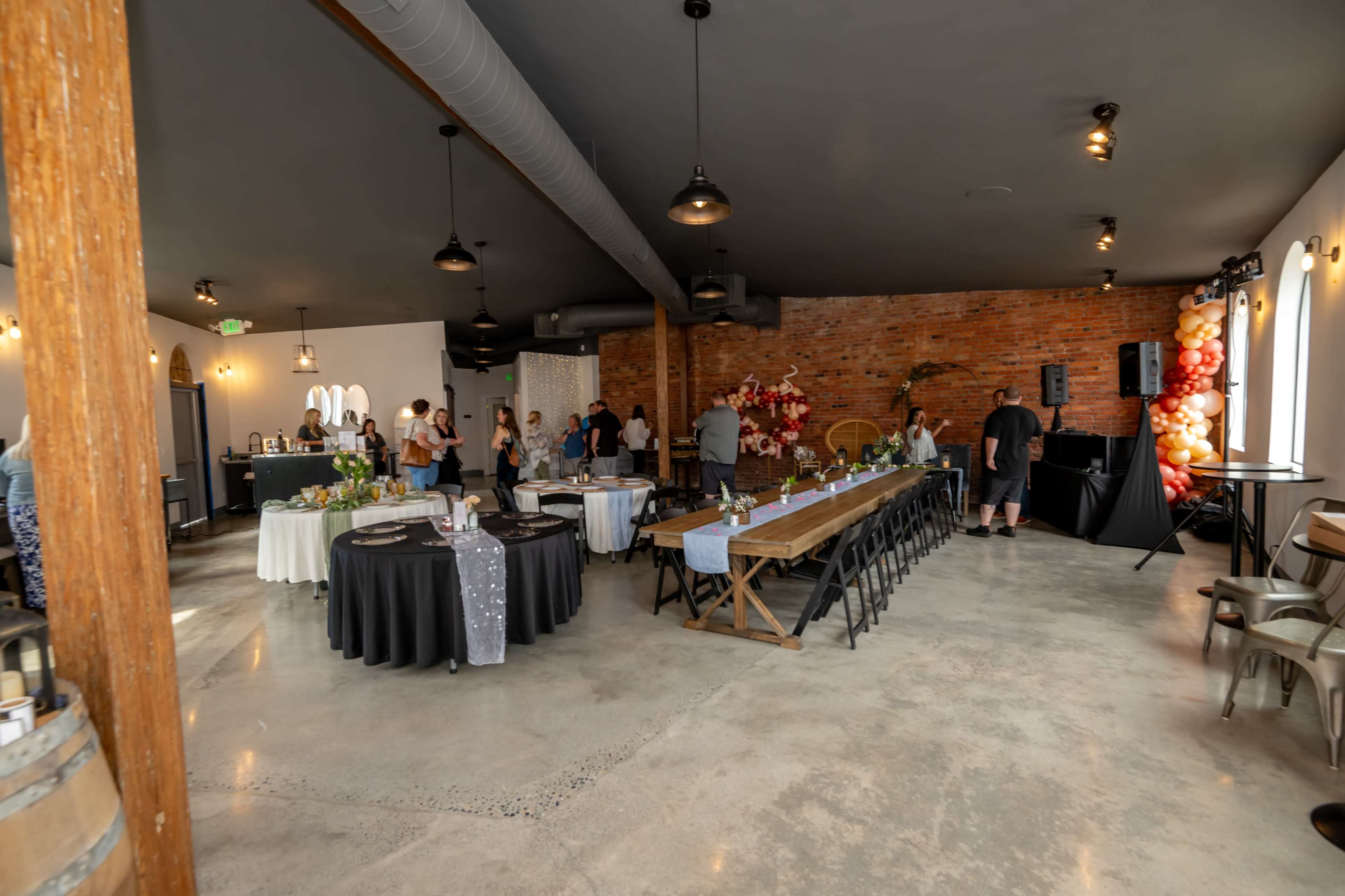 An event space with guests mingling, featuring a long wooden table, several round tables, and decorative elements against a brick wall.