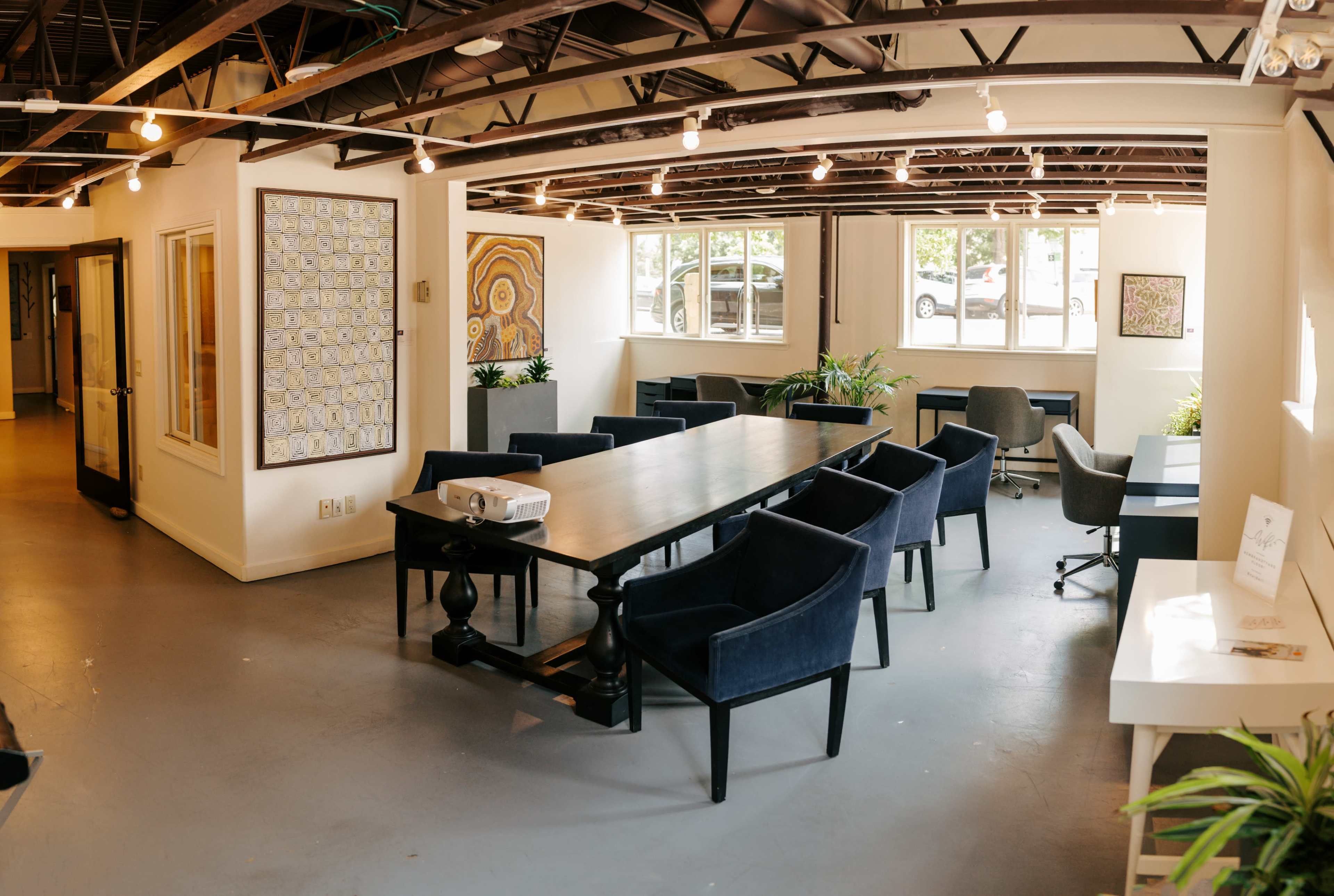 A spacious, well-lit office features a long table surrounded by dark chairs, with plants and artwork on the walls.