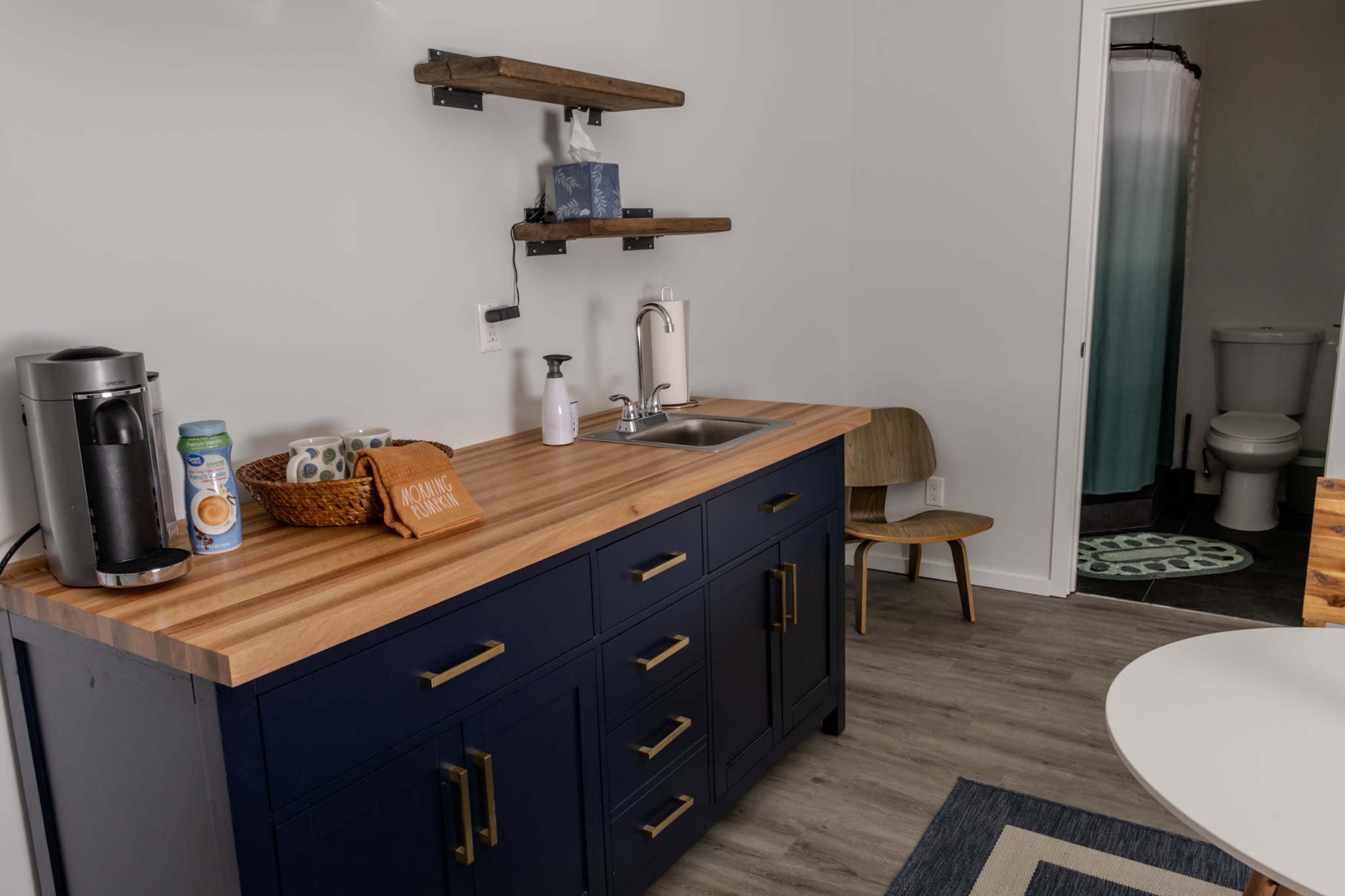 A compact kitchen area features a wooden countertop with a sink, dark blue cabinets, and shelves above, alongside a seating area and a bathroom visible in the background.